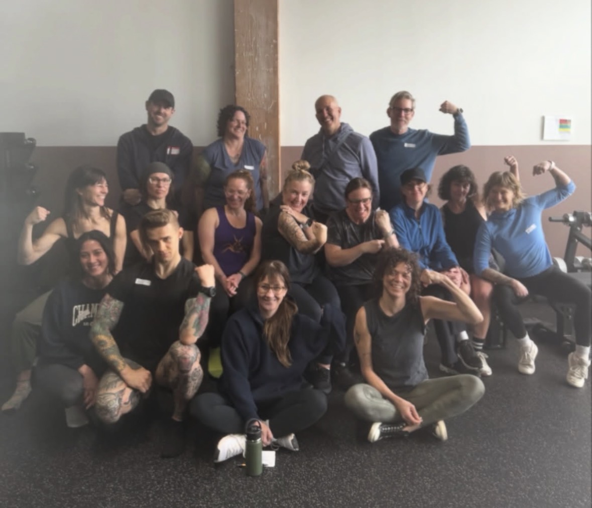 Group of 14 people posing indoors, some flexing muscles, some sitting or standing, smiling, with a wooden beam in the background.