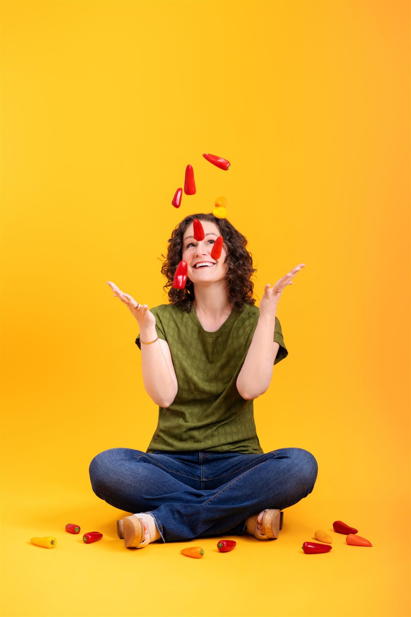 Person sitting cross-legged on yellow background, juggling red objects, smiling, with arms raised and scattered objects around.