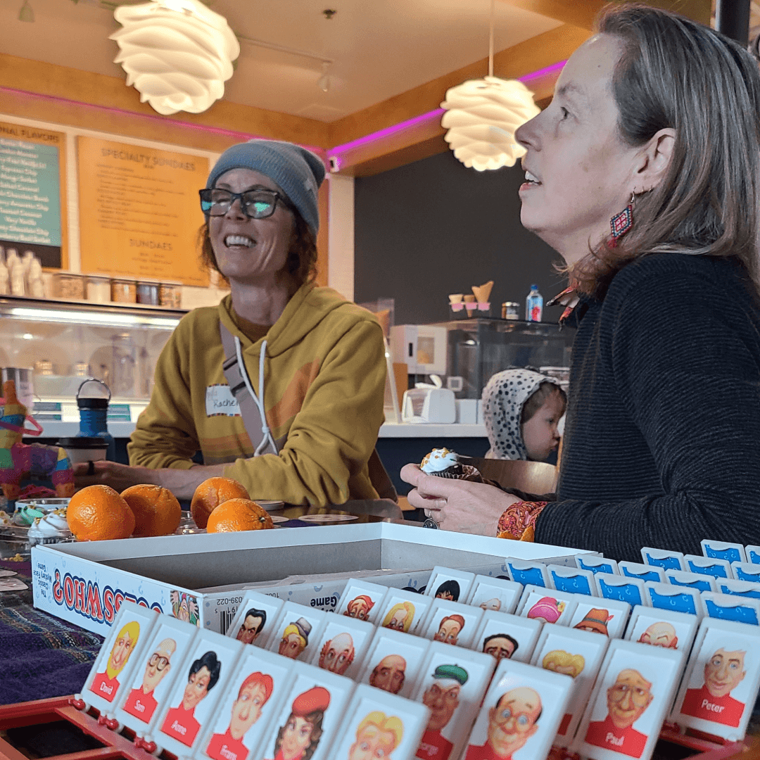 Two women talking in a cafe, one wearing glasses and a beanie, the other with long hair, with oranges in a box in front.