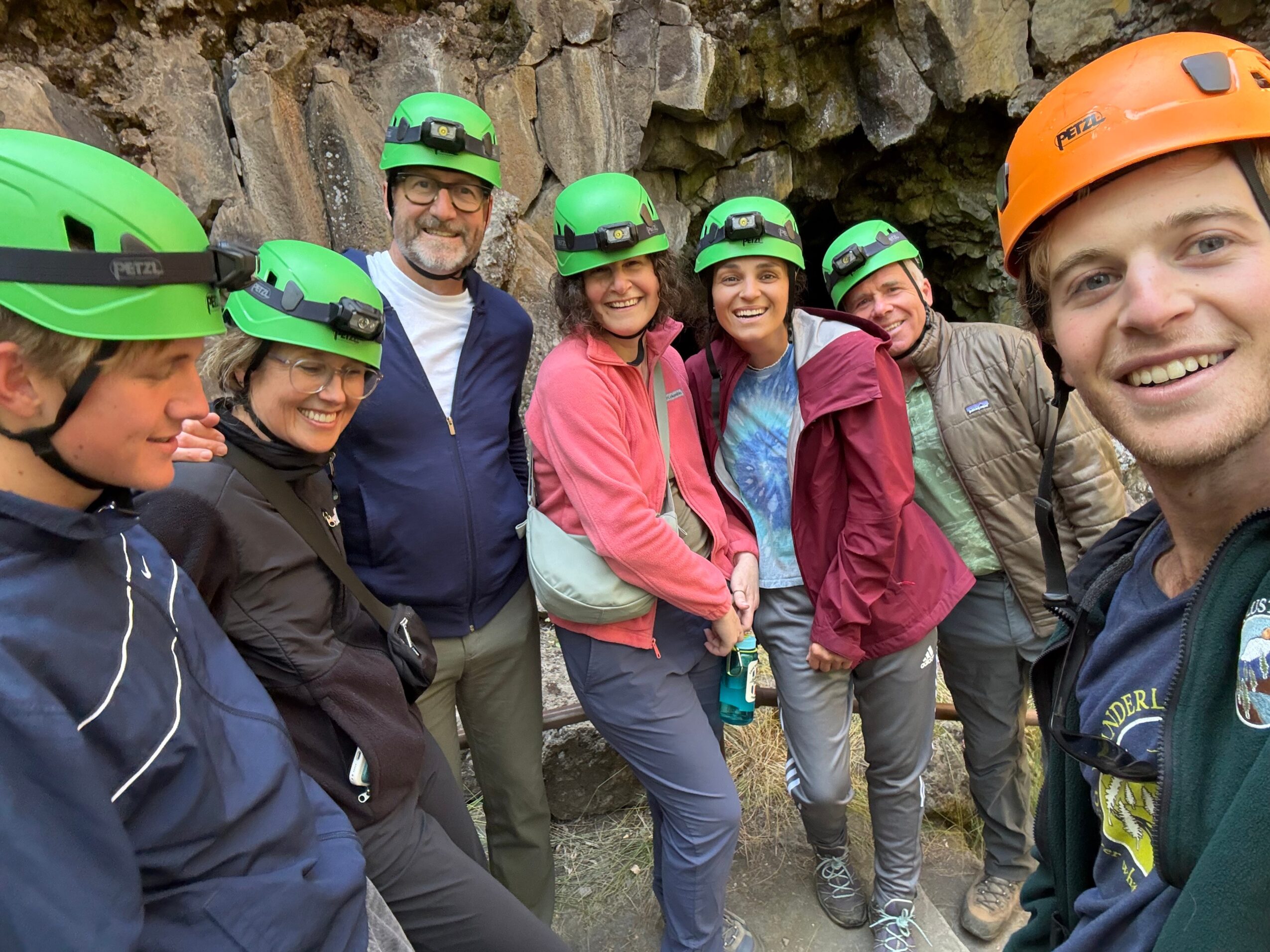 Group of seven people wearing helmets and outdoor clothing, standing outdoors near rocks, smiling for a photo.