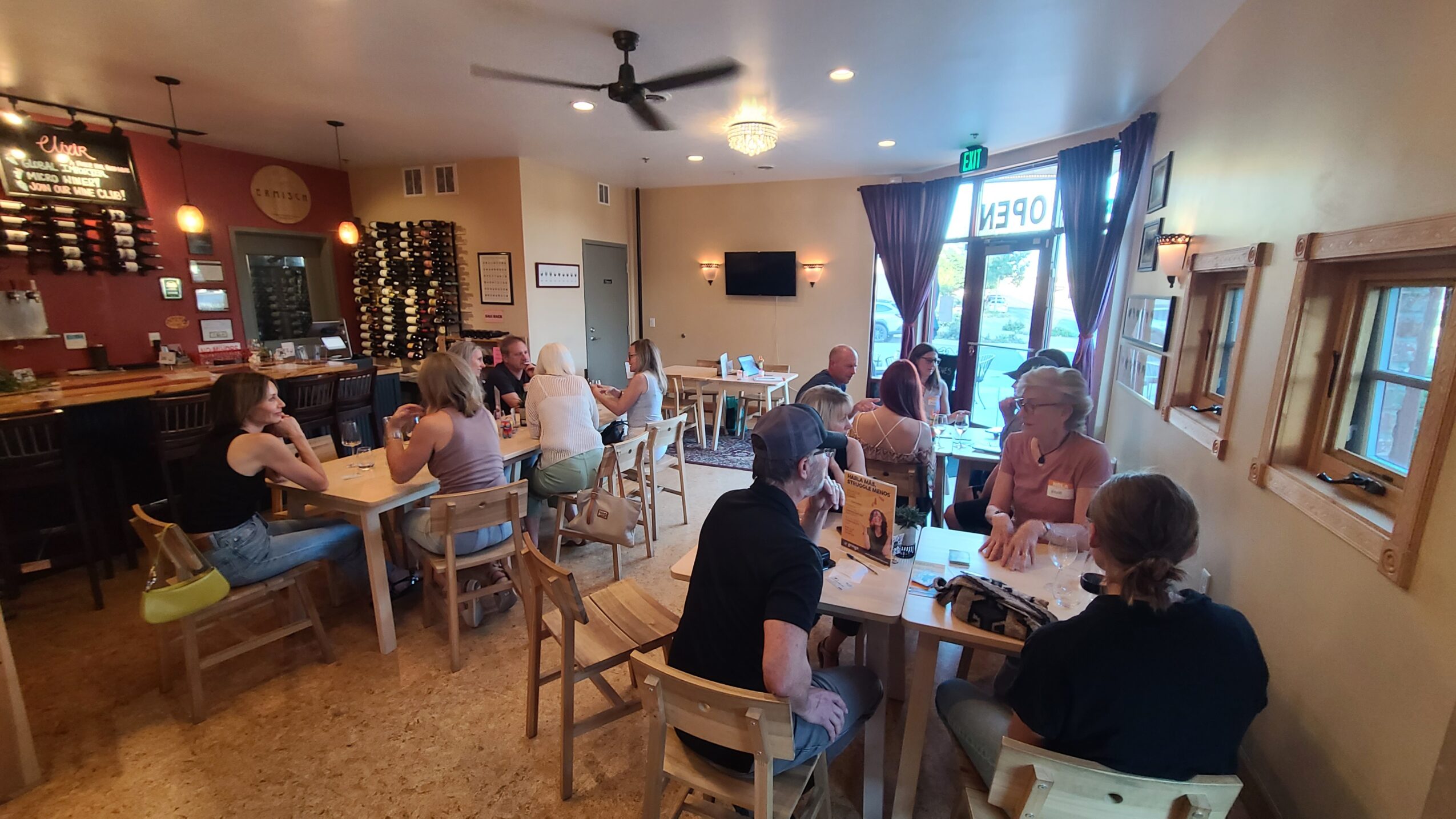 People sitting at tables inside a cafe, some near windows, with a bar area in the background, and a TV on the wall.