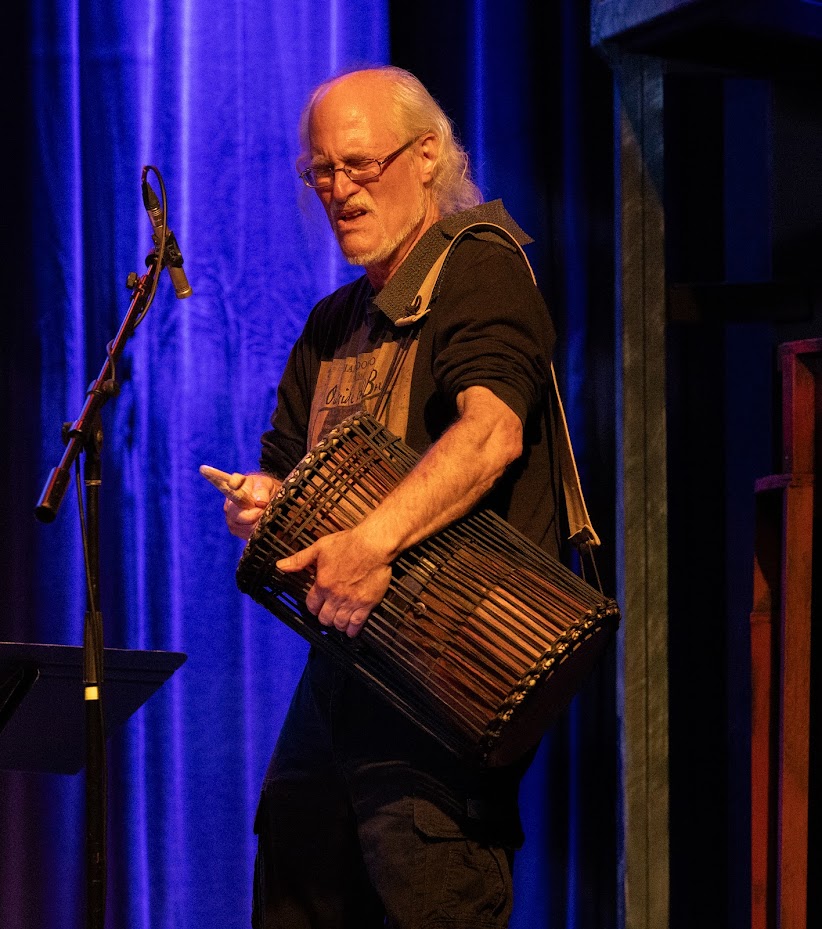 Older man with glasses playing a large drum on stage with blue curtains in the background.