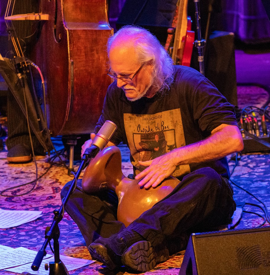 Older man with long white hair and glasses playing a drum with a microphone nearby, seated on a stage with musical instruments.