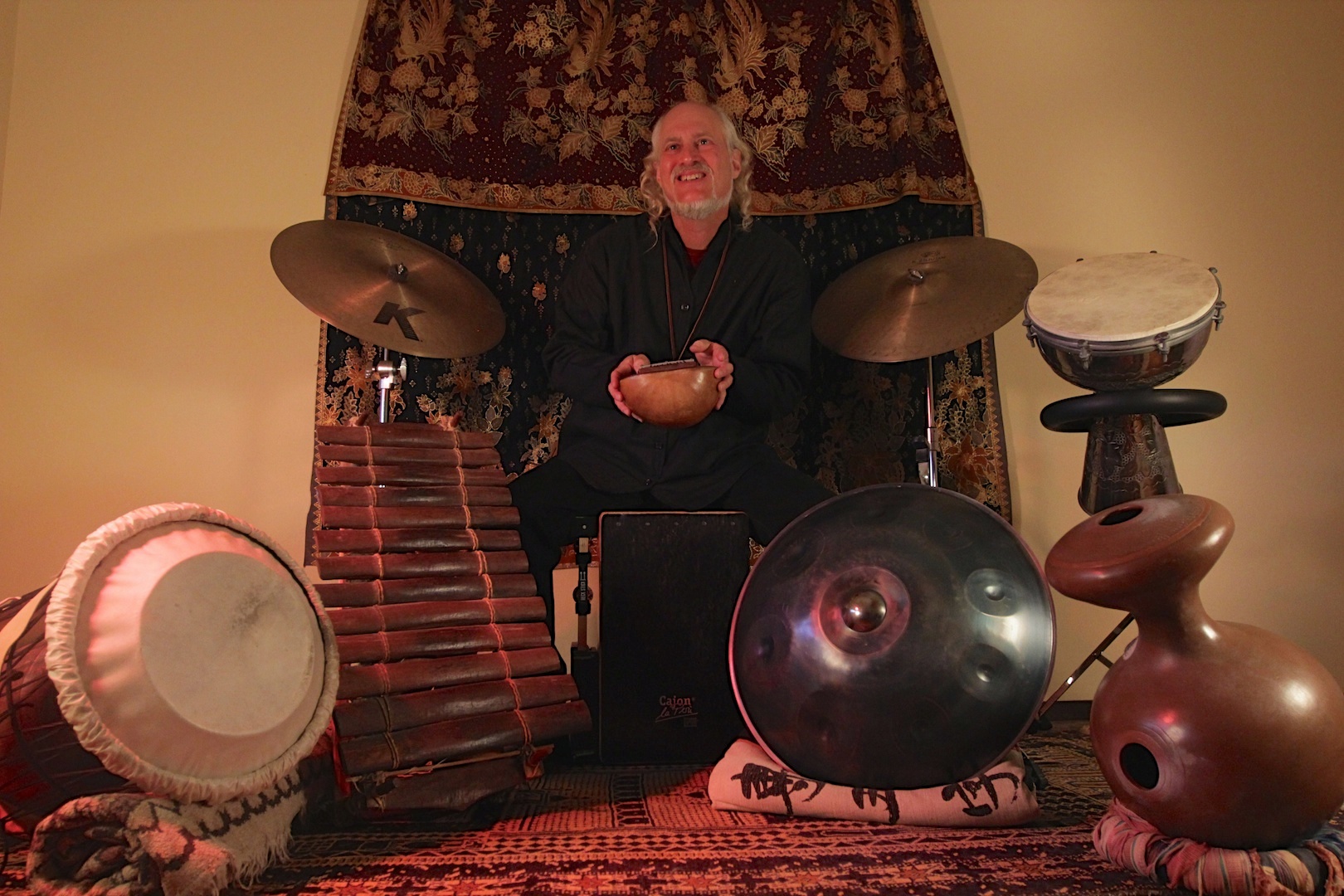 Person sitting with musical instruments, including drums and percussion, in a room with a curtain backdrop.