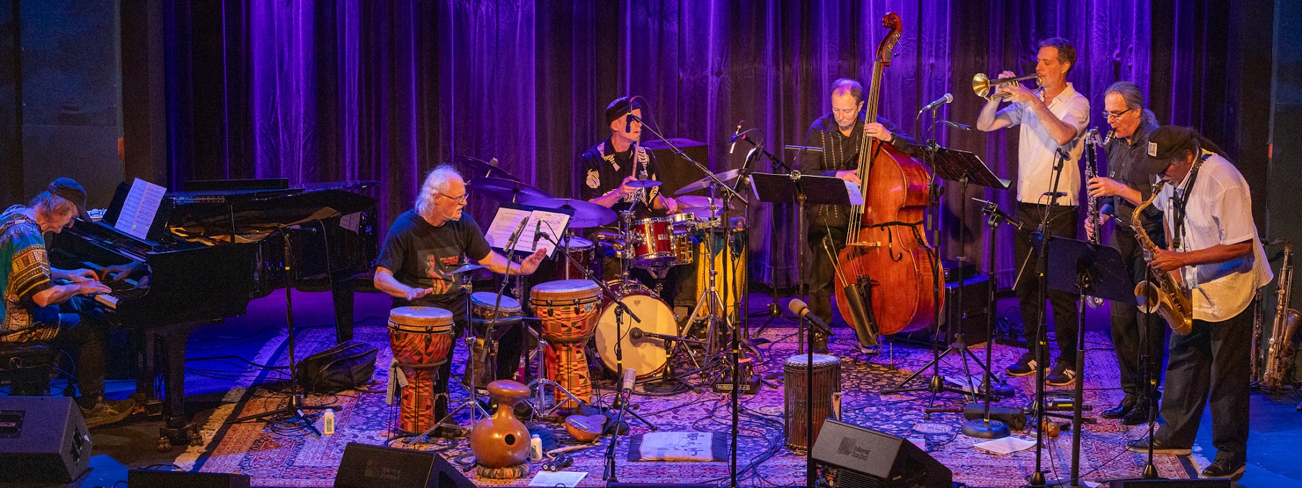 Musicians perform on stage with instruments, purple lighting, and a dark curtain backdrop.