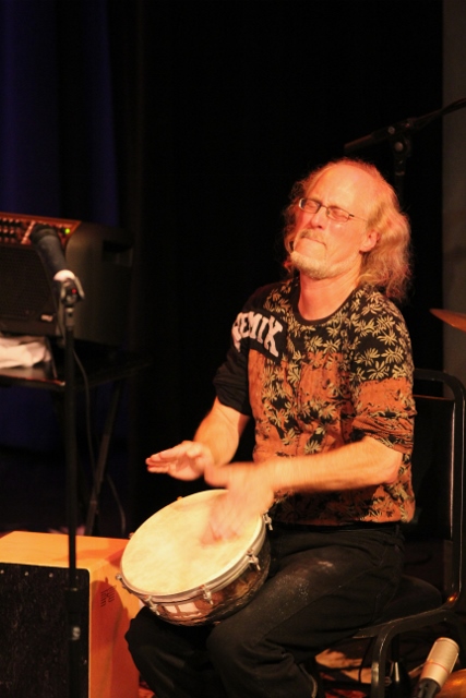 Person with glasses and long hair playing a drum on stage, wearing a patterned shirt, with dark background.
