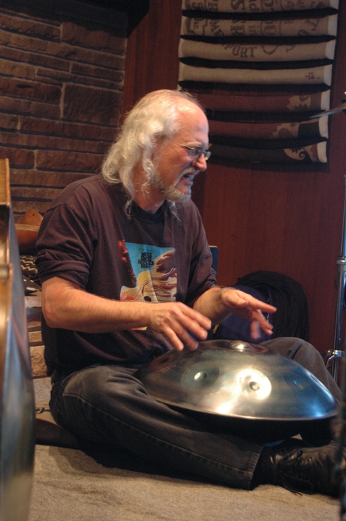 Older man with long white hair and beard playing a handpan drum, sitting cross-legged indoors.