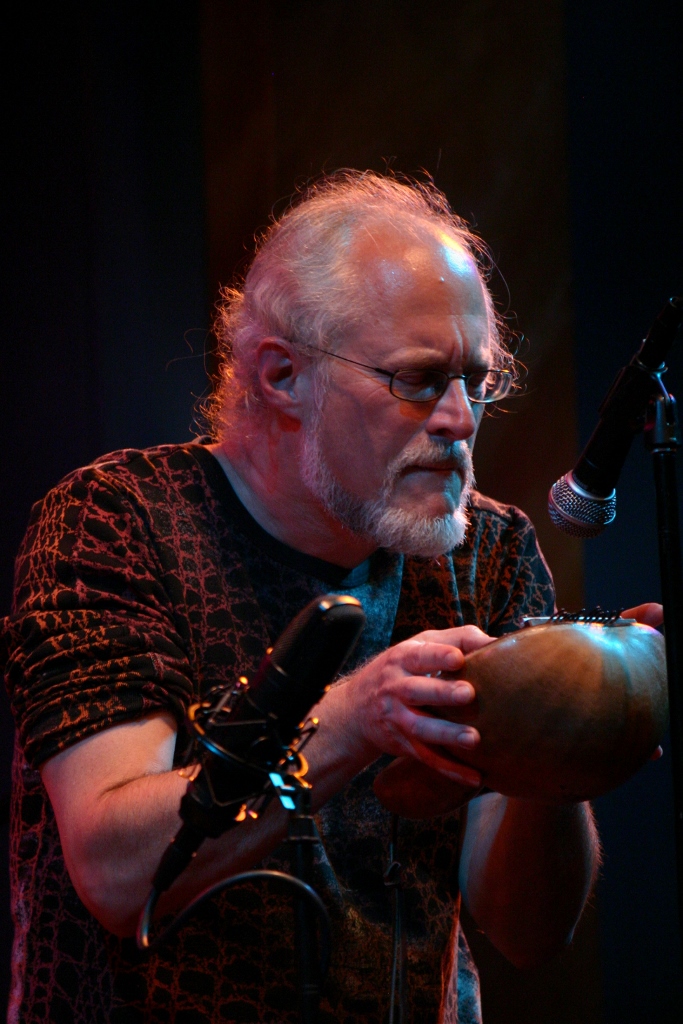 Elderly man with glasses and white hair playing a percussion instrument on stage, with a microphone nearby.