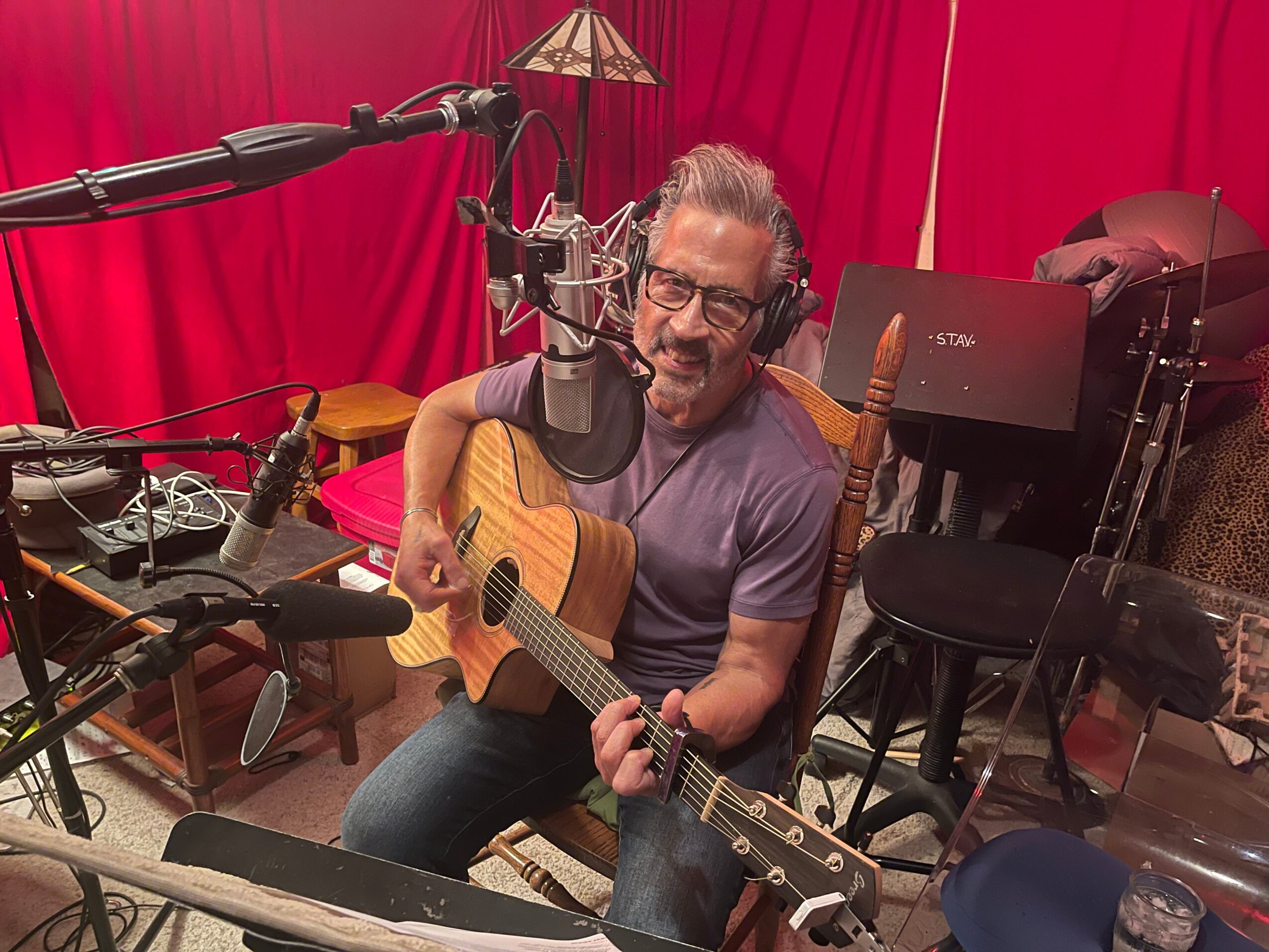 Man with glasses and headphones playing acoustic guitar in recording studio with red curtains and musical equipment