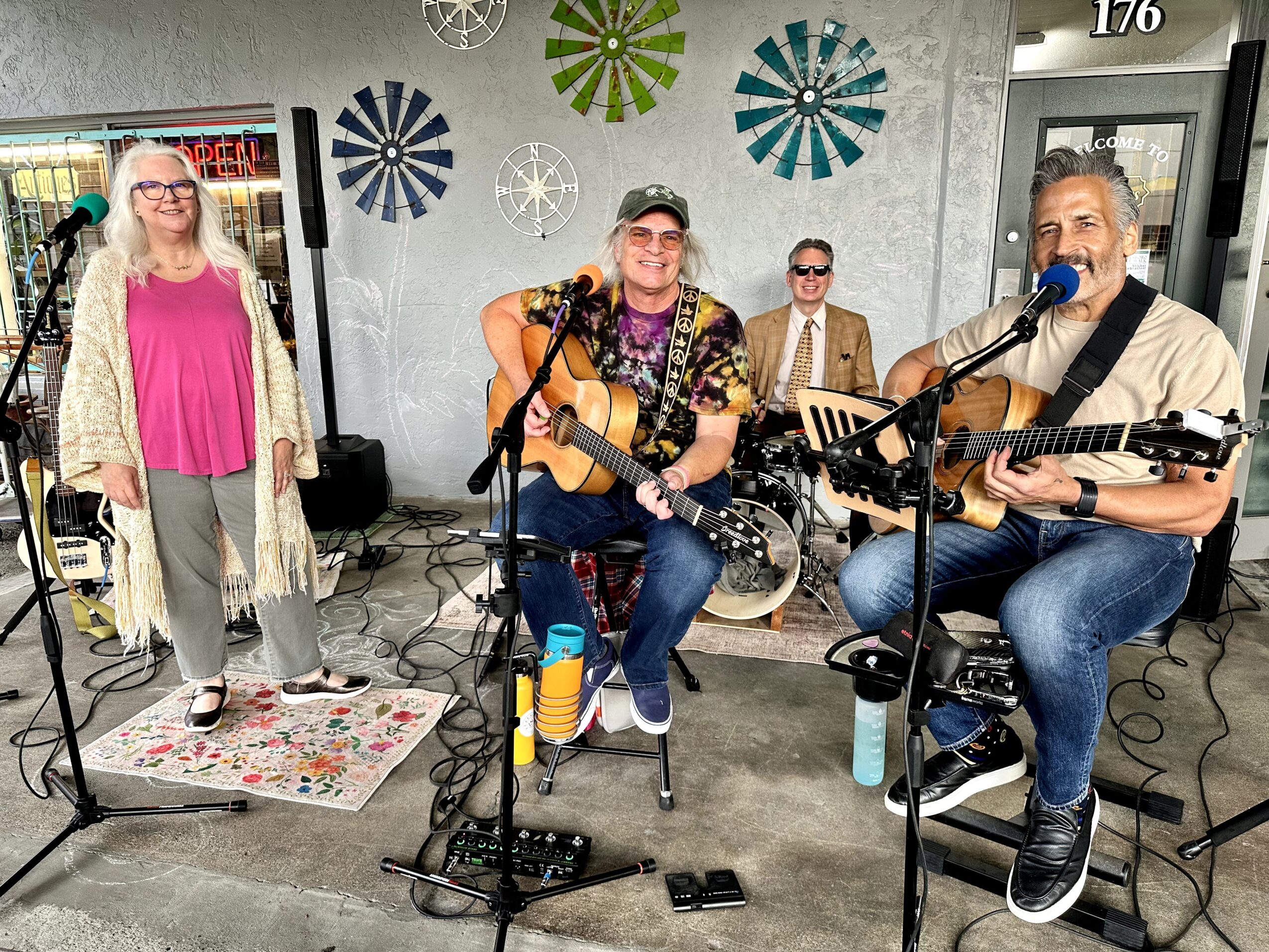 Four people performing music indoors, two playing guitars and singing, one woman standing nearby, colorful wall decorations, casual setting.