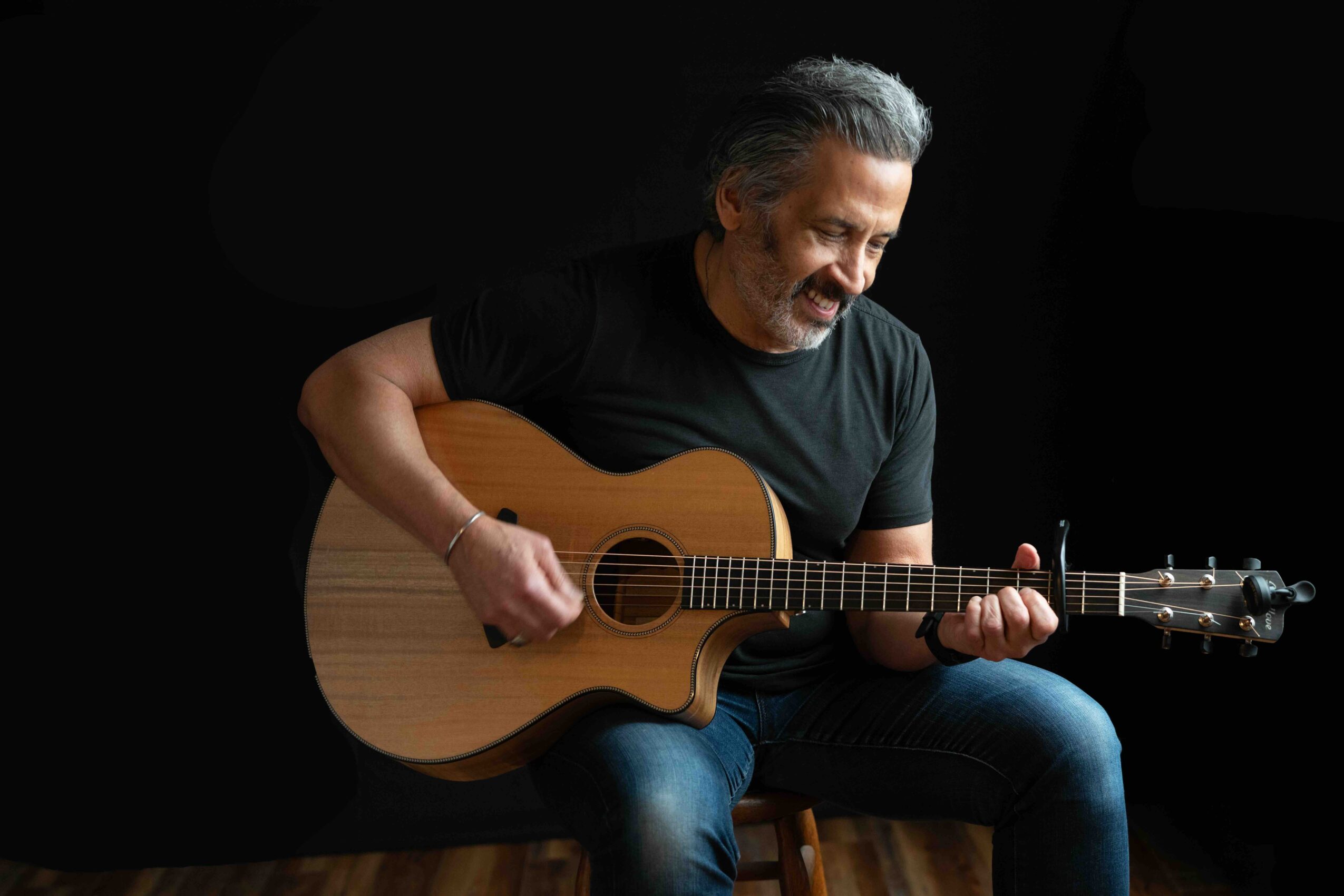 Man with gray hair playing an acoustic guitar, smiling, against a black background.