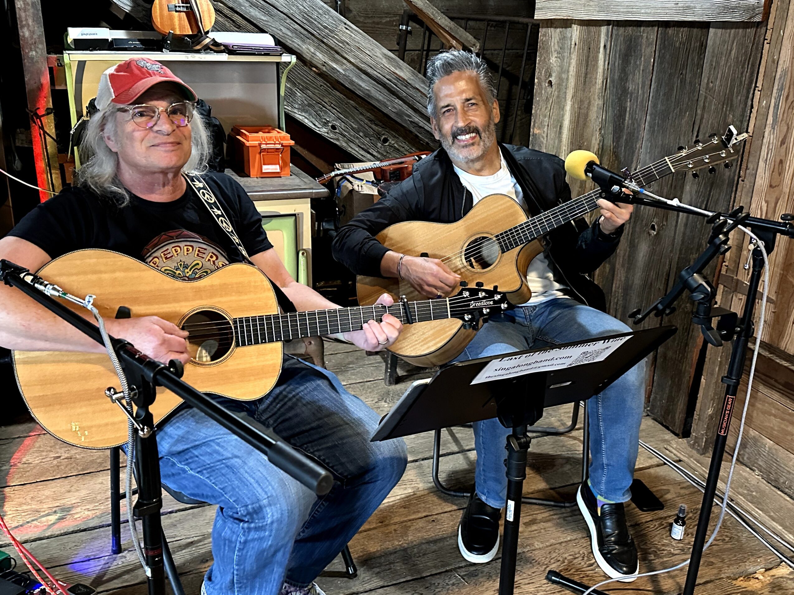 Two men playing acoustic guitars and singing, seated indoors with musical equipment and wooden walls.