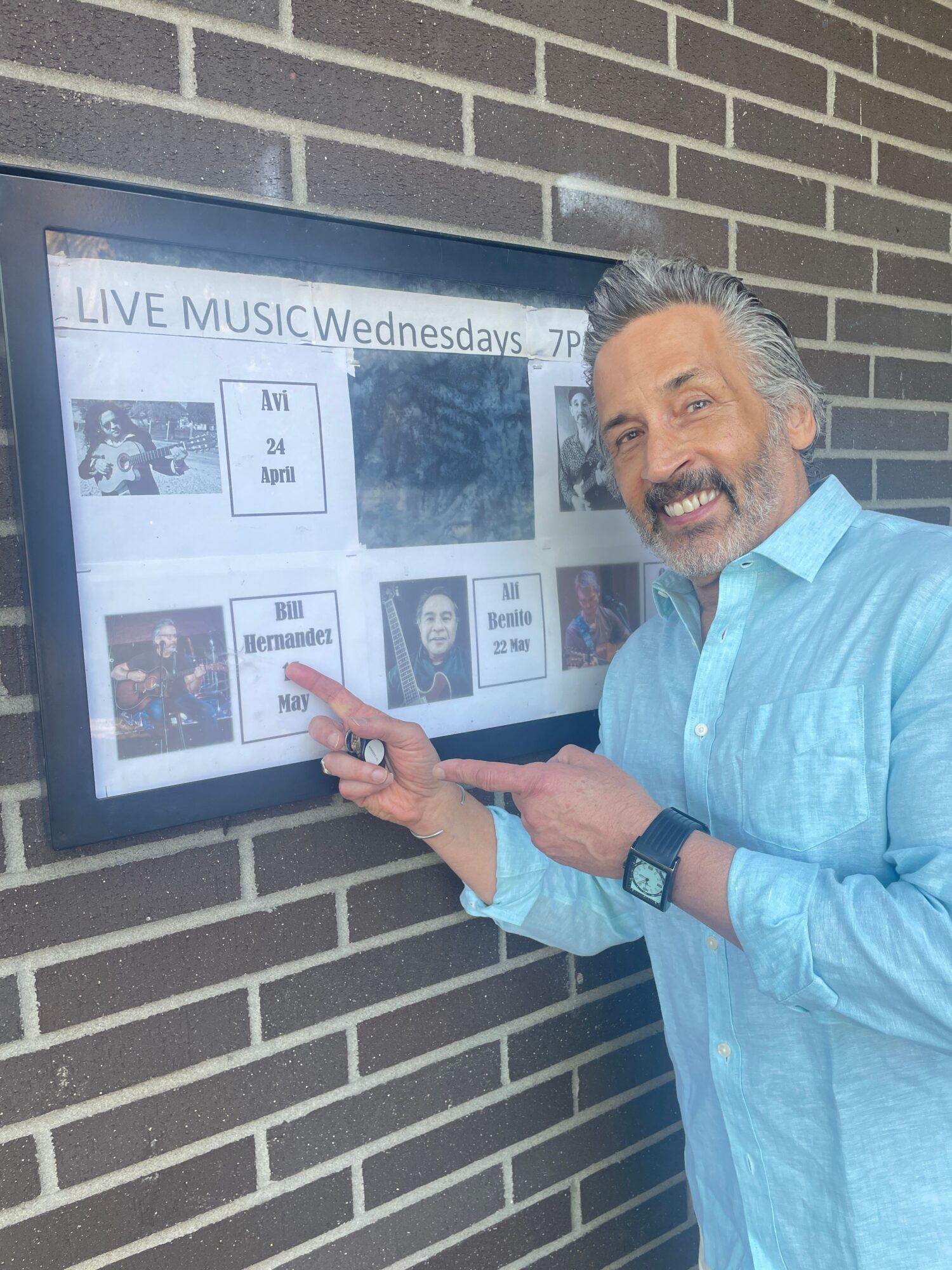 Person with gray hair and beard smiling, pointing at a large screen display, standing against a brick wall.