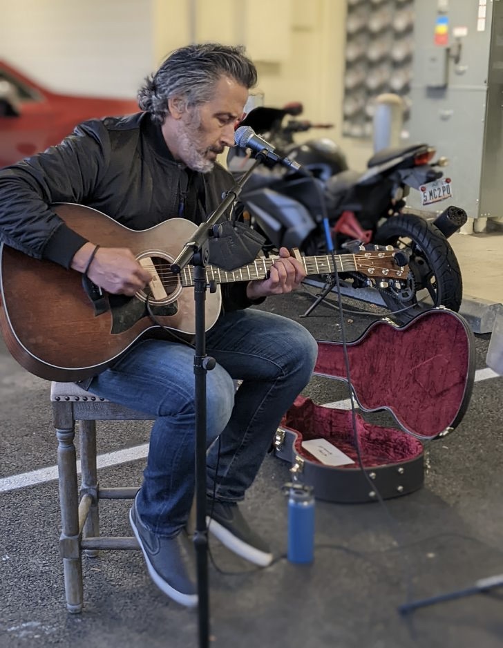 Man playing guitar on a chair, with an open guitar case and motorcycles in the background.