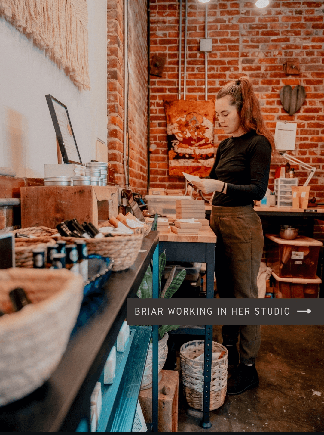 Woman browsing products in a brick-walled studio with art and supplies, standing at a table, holding a book or catalog.
