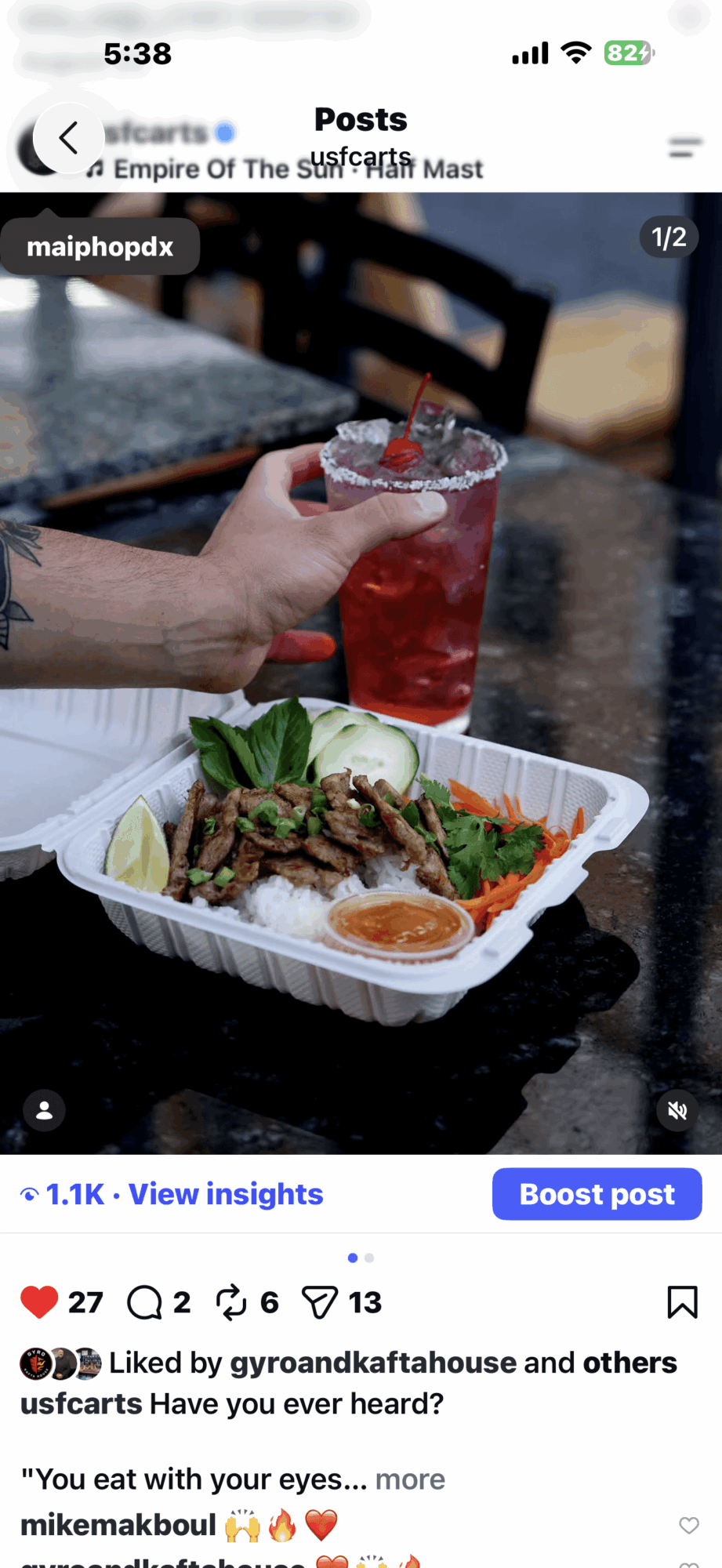 Hand holding a glass of red drink with ice, in front of a food container with grilled meat, vegetables, and sauce on a dark table.