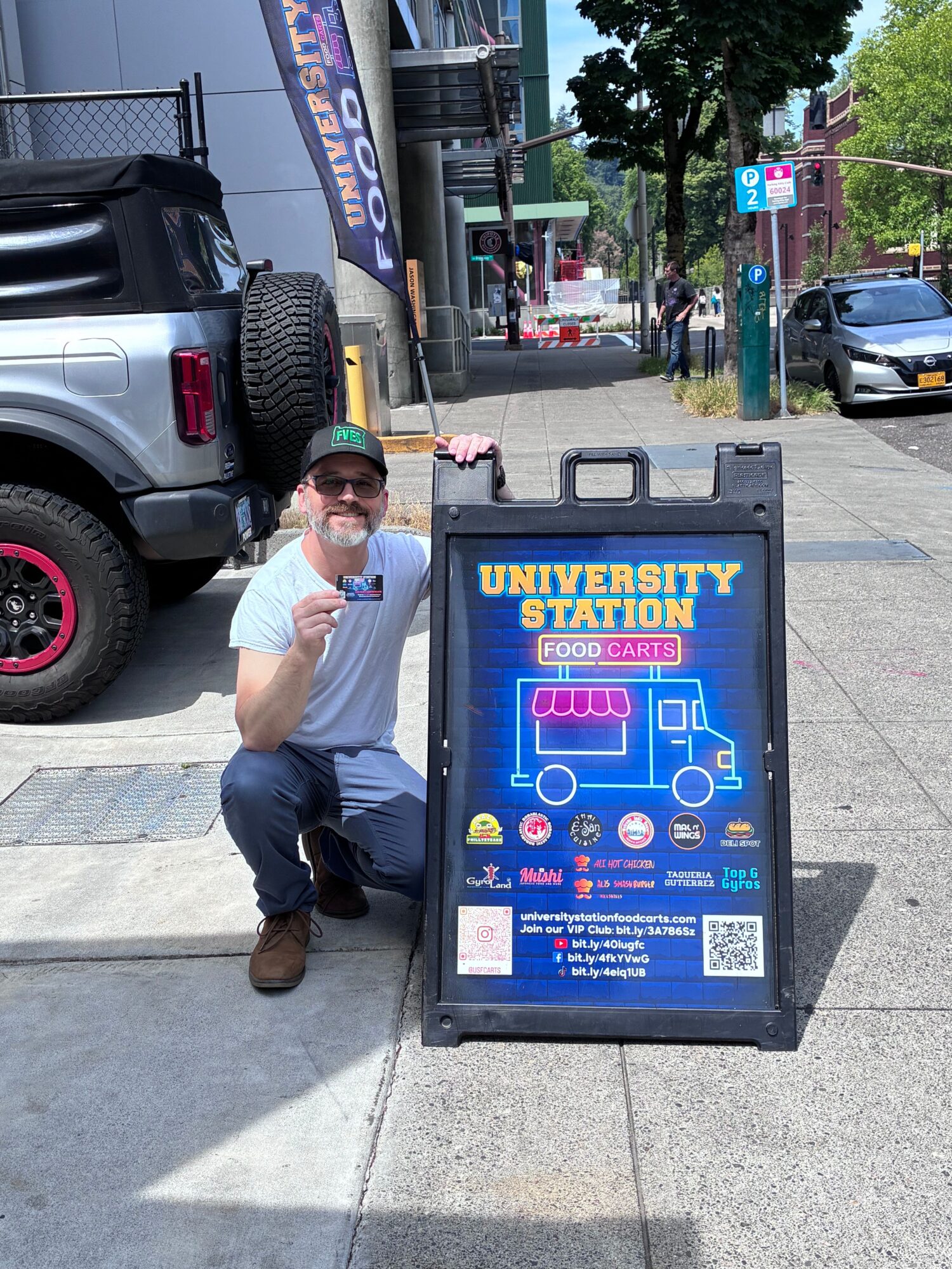 Person taking a selfie next to a car and a university station sign on a city sidewalk.