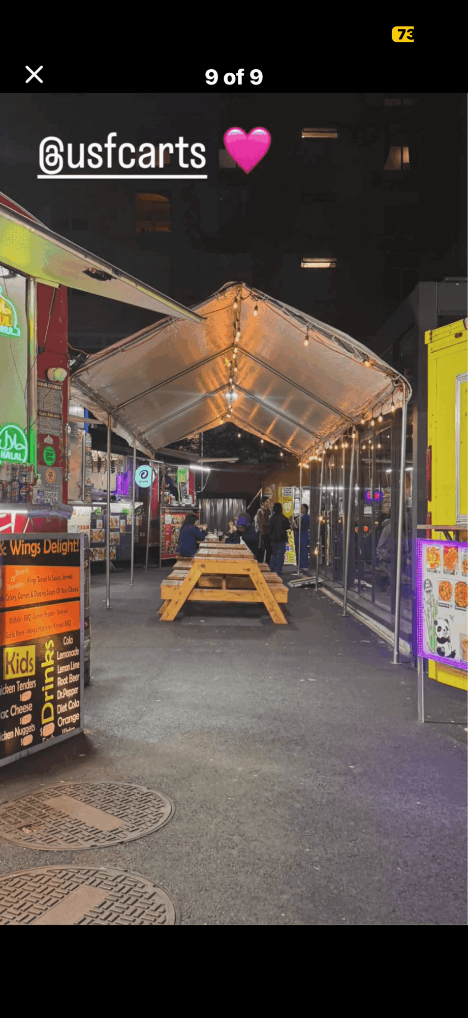 Empty street with a canopy, benches, and colorful signs at night, illuminated by streetlights and neon lights.