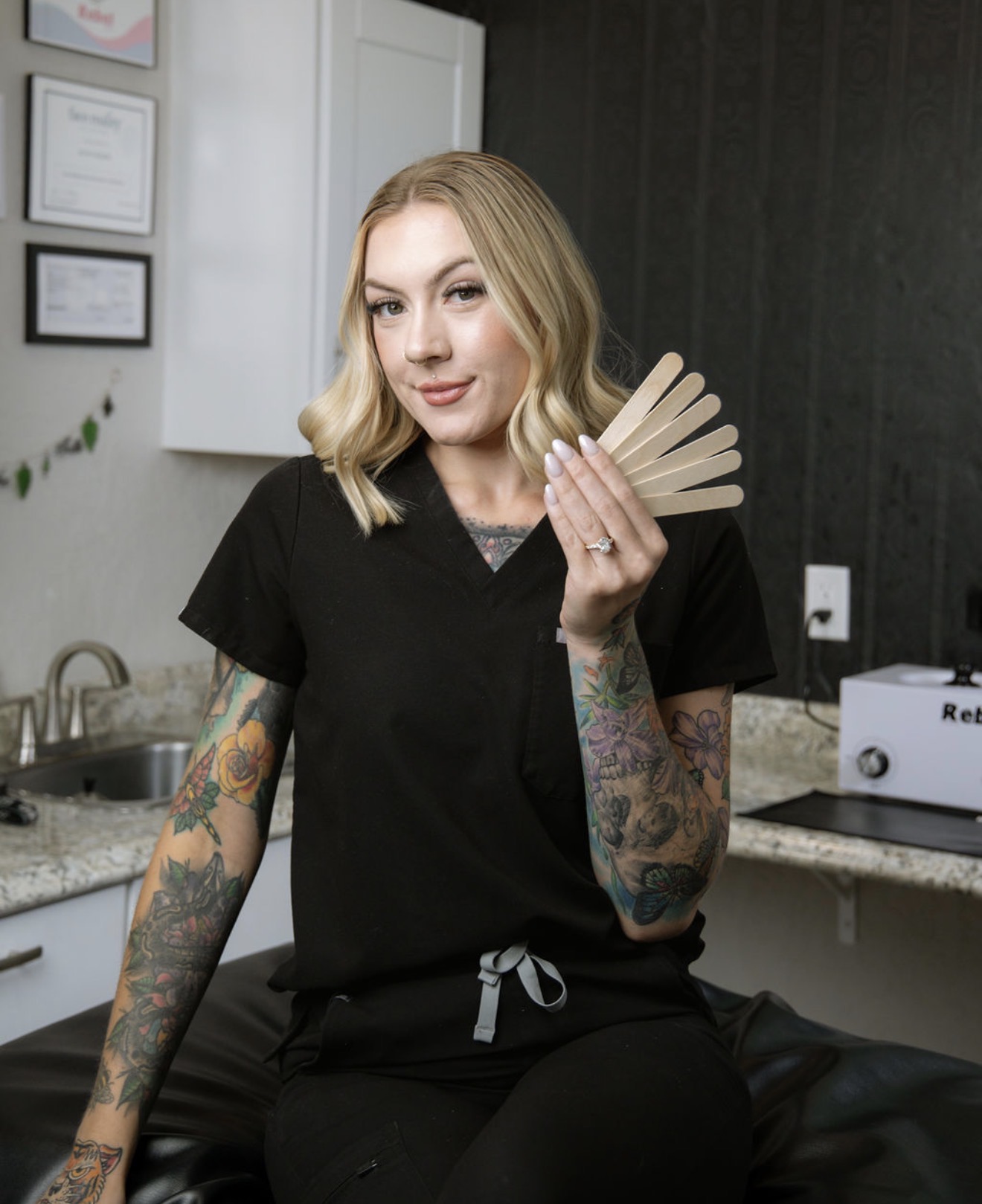 Young woman with blonde hair and tattoos, wearing black scrubs, holding a fan of cards, standing in a kitchen.