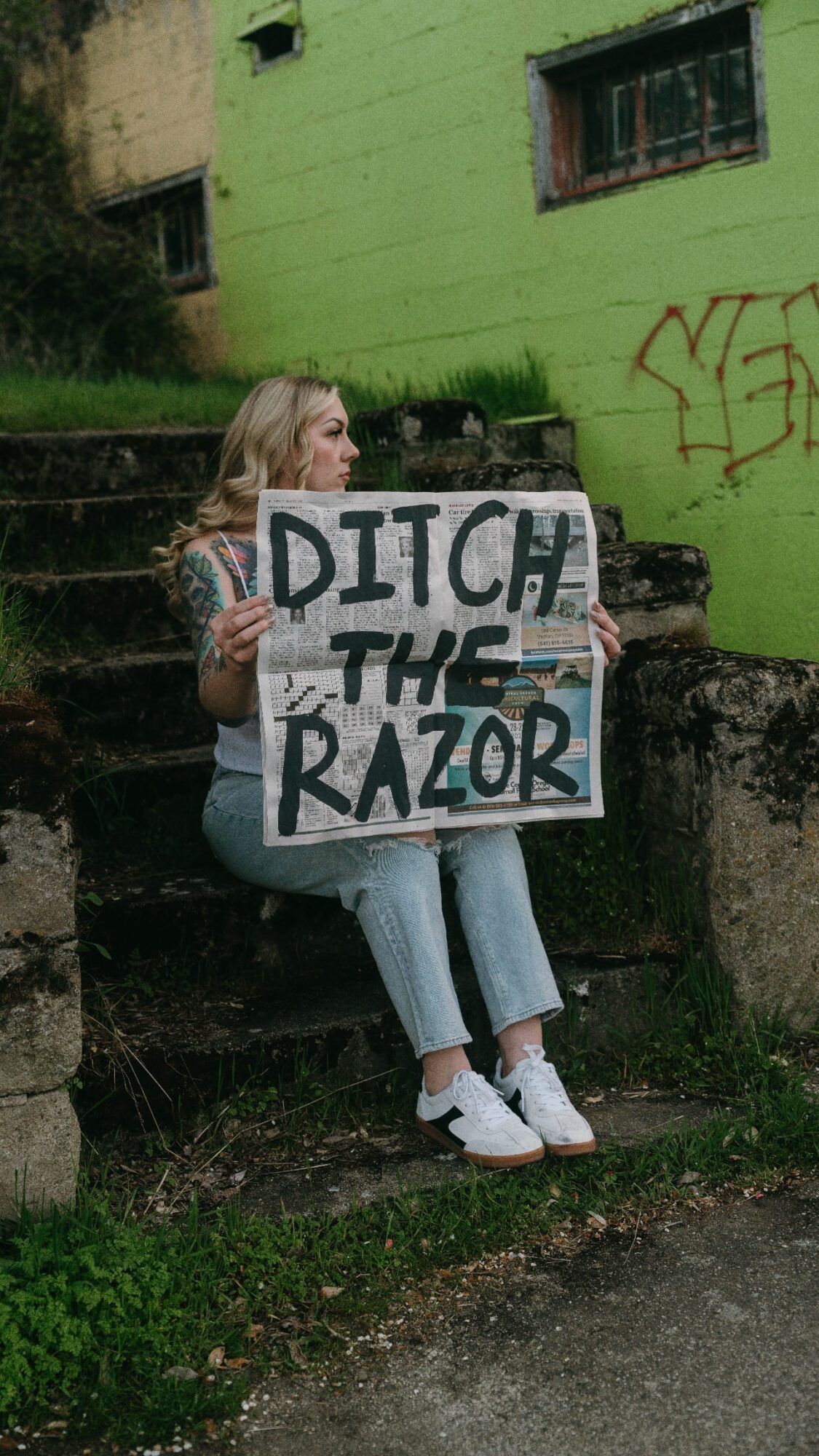 Person sitting on steps holding a sign that says 'Ditch the Razor' in front of a green building with graffiti.