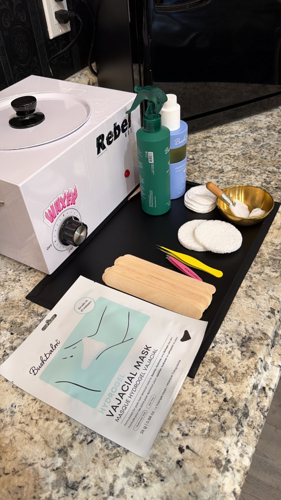 Skincare products, wooden spatula, and a mask packet on a black tray on a granite countertop.