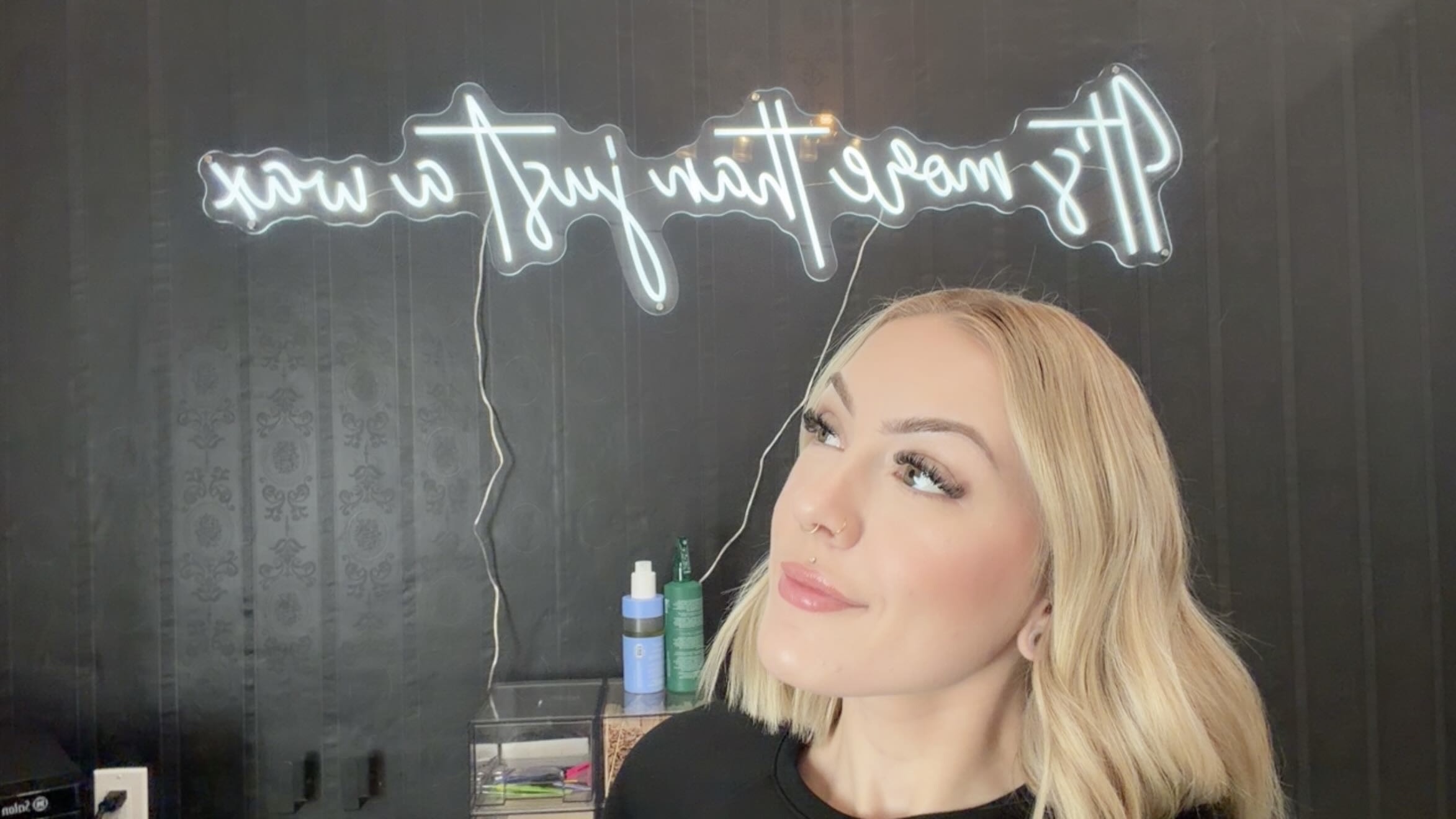 Woman with blonde hair looking to the side in front of a neon sign, with bottles and a mirror nearby.