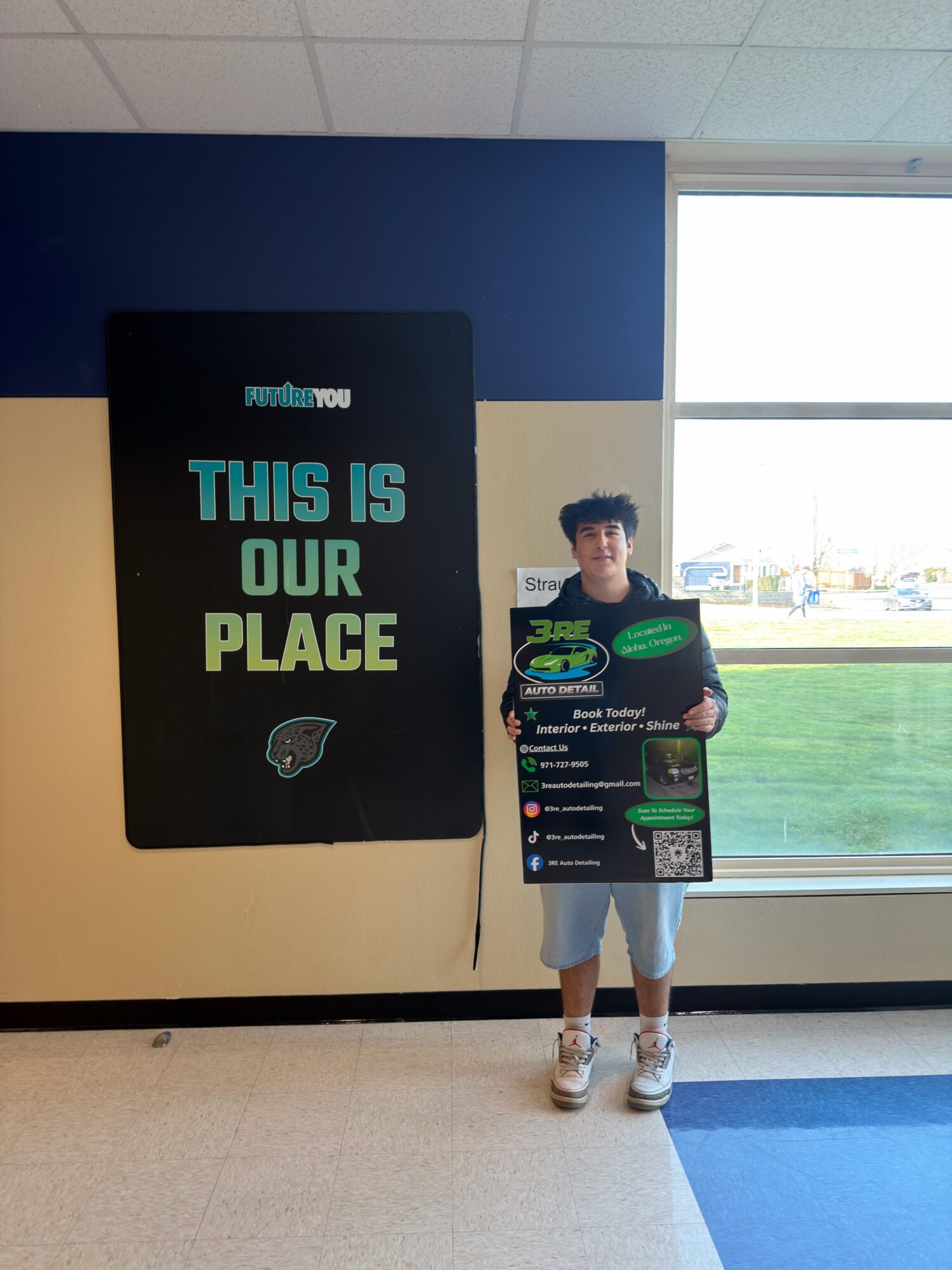 Young person holding a promotional sign in front of a large window and a sign that reads 'This is our place'.