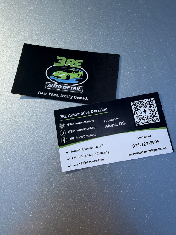Black business card with green and white logo, QR code, and contact information, tilted sideways on a gray surface.