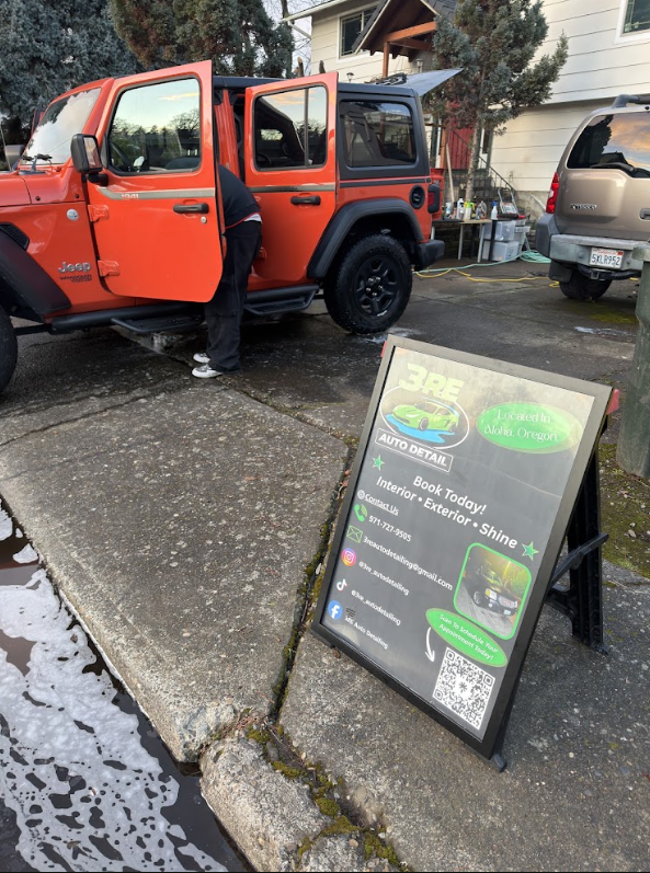 Red and black Jeep parked on driveway with open door, person standing nearby, and a signboard advertising vehicle services, with snow on ground.