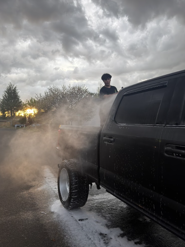 Person standing near a black pickup truck with dust or water spray, cloudy sky overhead, trees in background.