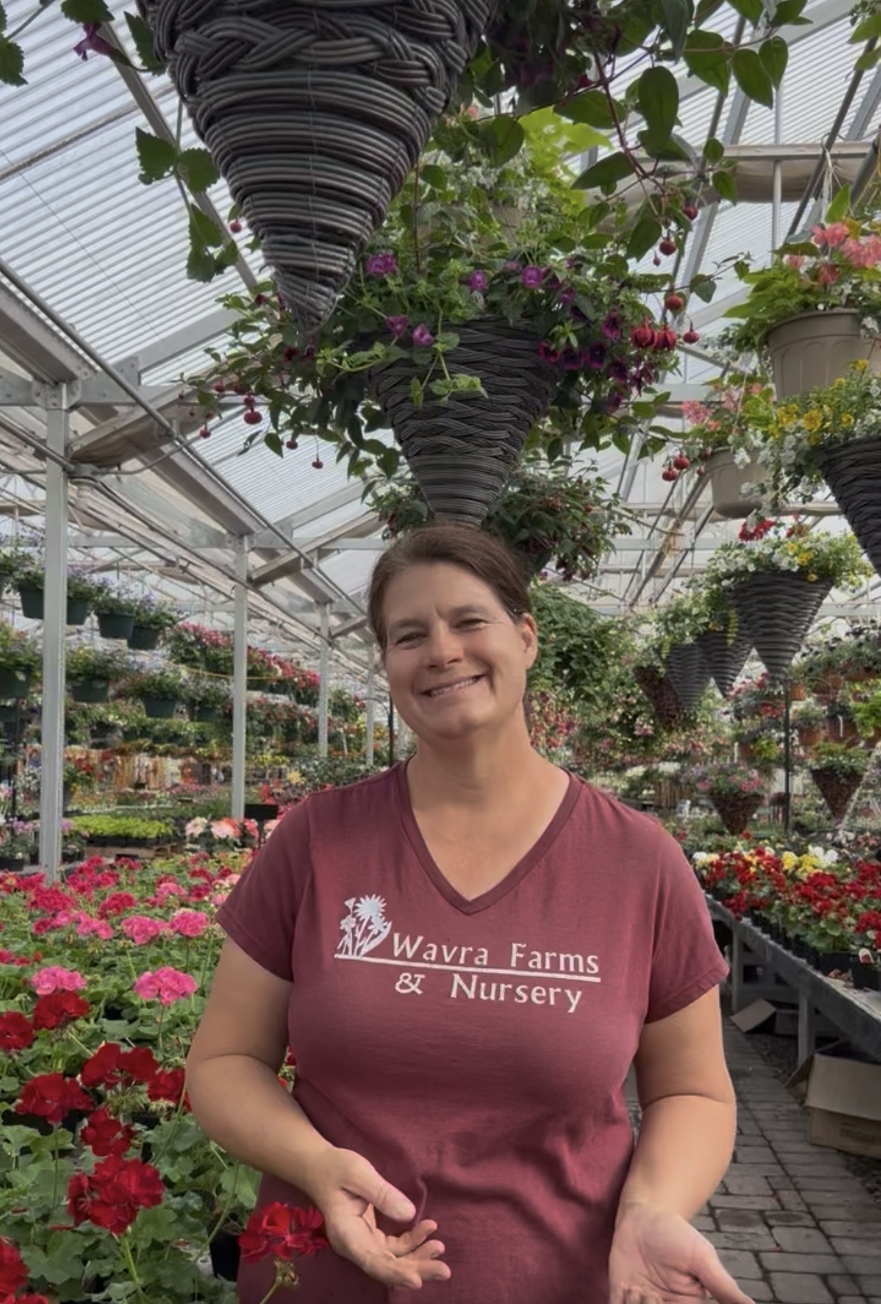 Smiling woman in a nursery surrounded by hanging and potted flowers in a greenhouse.