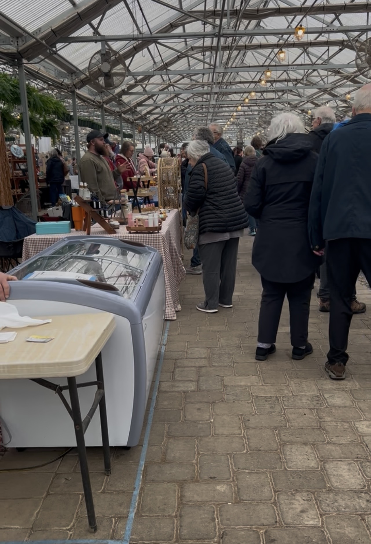 People browse market stalls inside a greenhouse with glass ceiling, some wearing coats and hats, with a table and a machine in foreground.