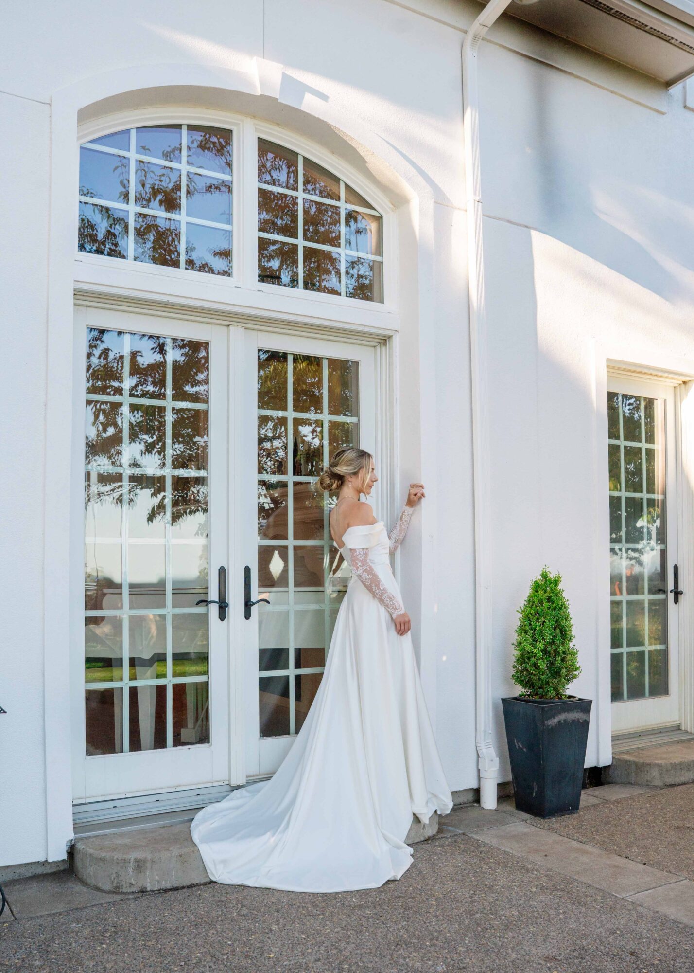 Woman in a white wedding dress standing outside near glass doors and a potted plant, with reflections of trees in the glass.
