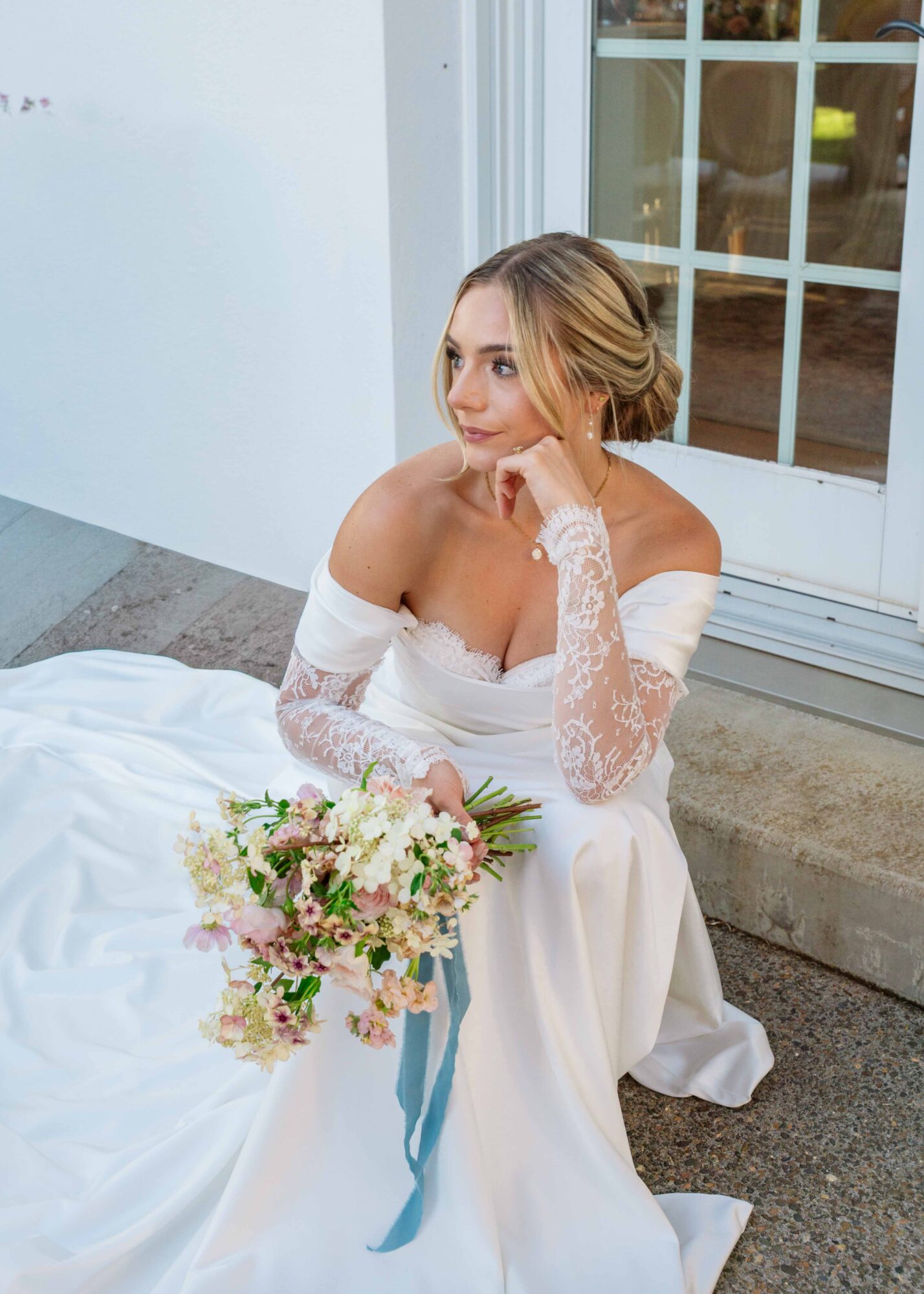 Woman in wedding dress sitting outside near a window, holding a bouquet of flowers, with her hand on her chin.