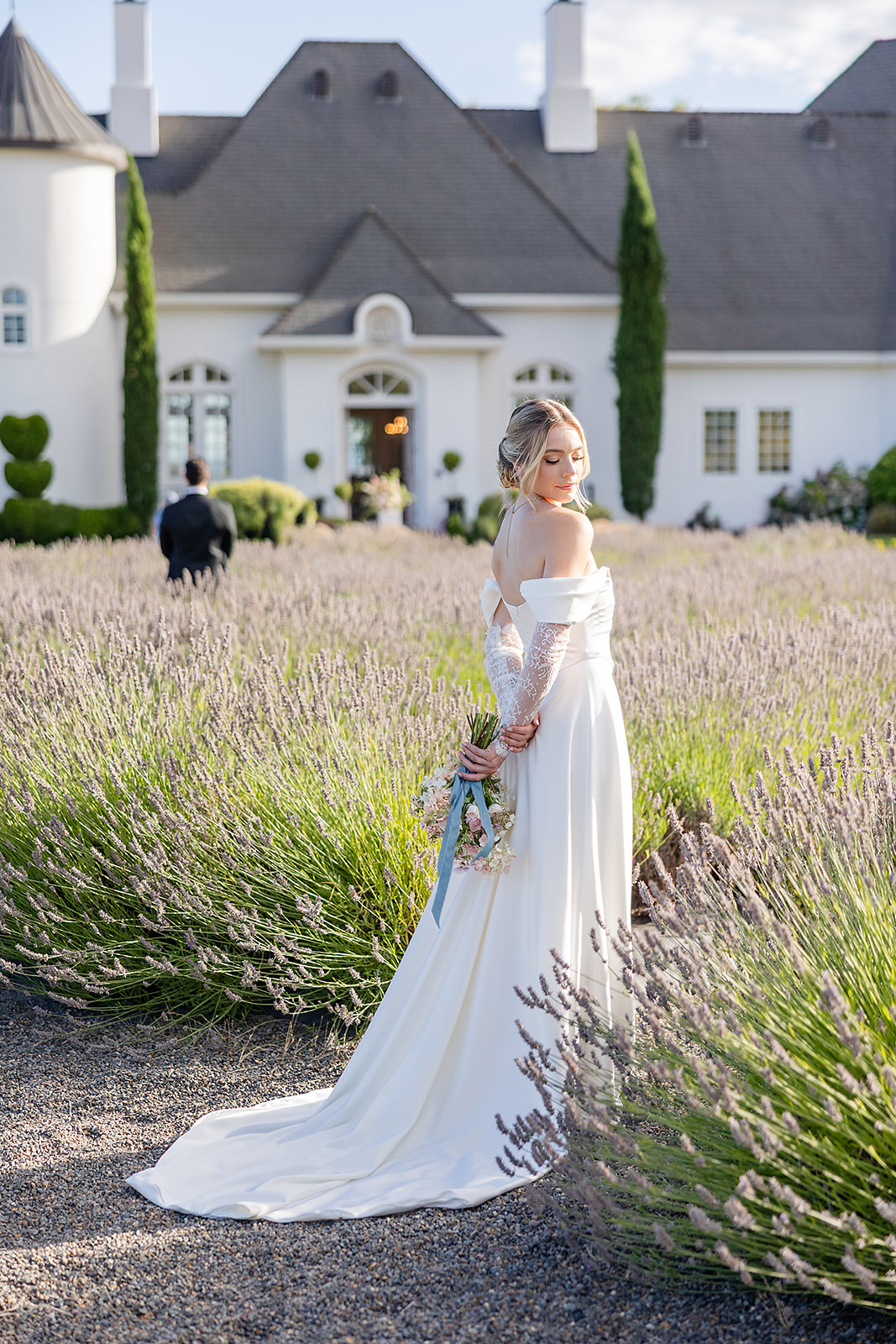 Woman in a white dress holding a bouquet in a lavender field with a large white house in the background.