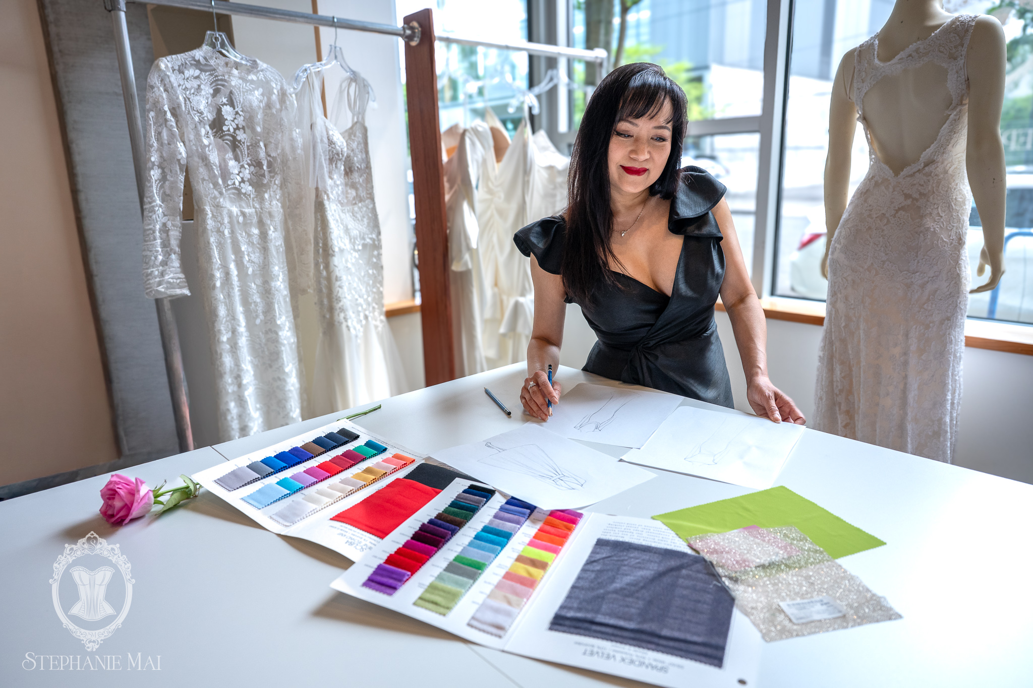 Woman sitting at a table with fabric swatches and sketches, surrounded by wedding dresses and mannequins in a bridal shop.