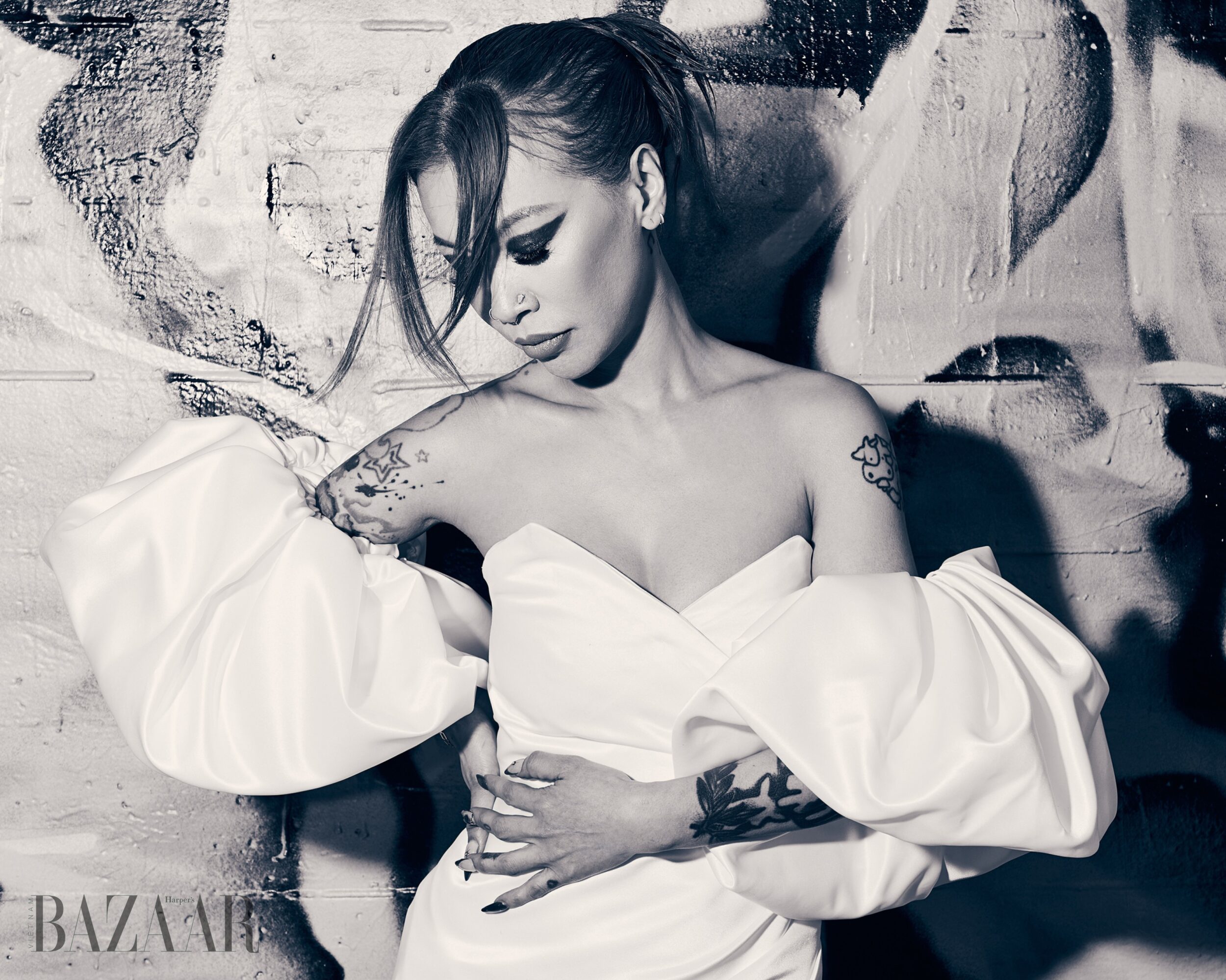 Woman with tattoos wearing an off-shoulder dress, looking down, against a textured wall background.