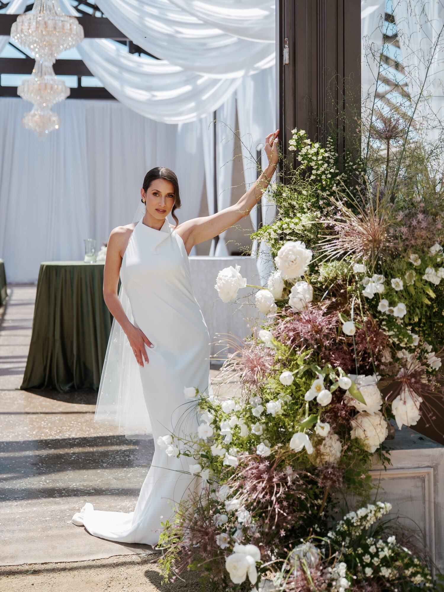 Woman in white dress standing next to large floral arrangement with white and pink flowers, indoors with draped ceiling and chandeliers.