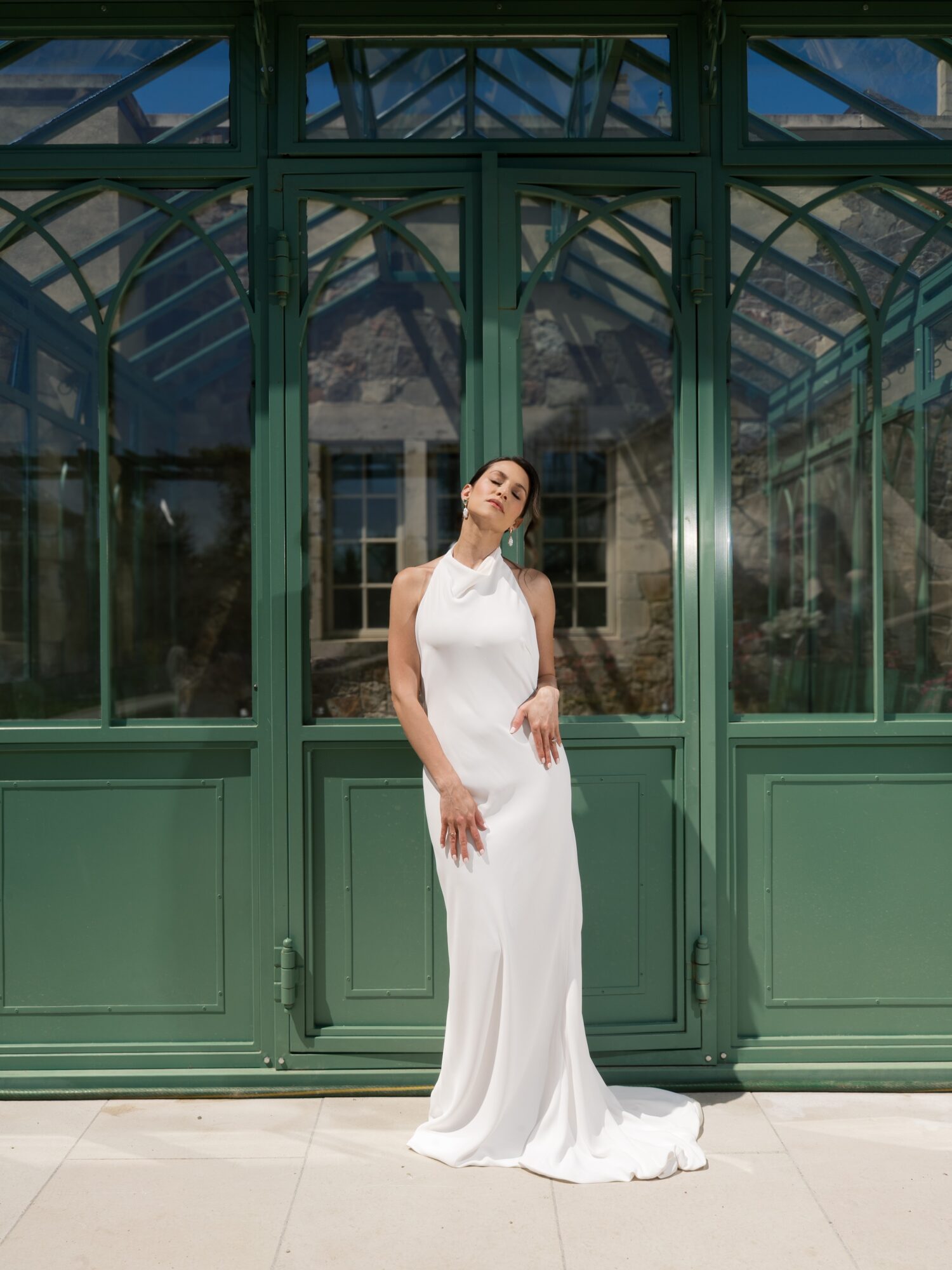 Woman in a white dress standing in front of a large green glass structure, sunlight shining on her face.