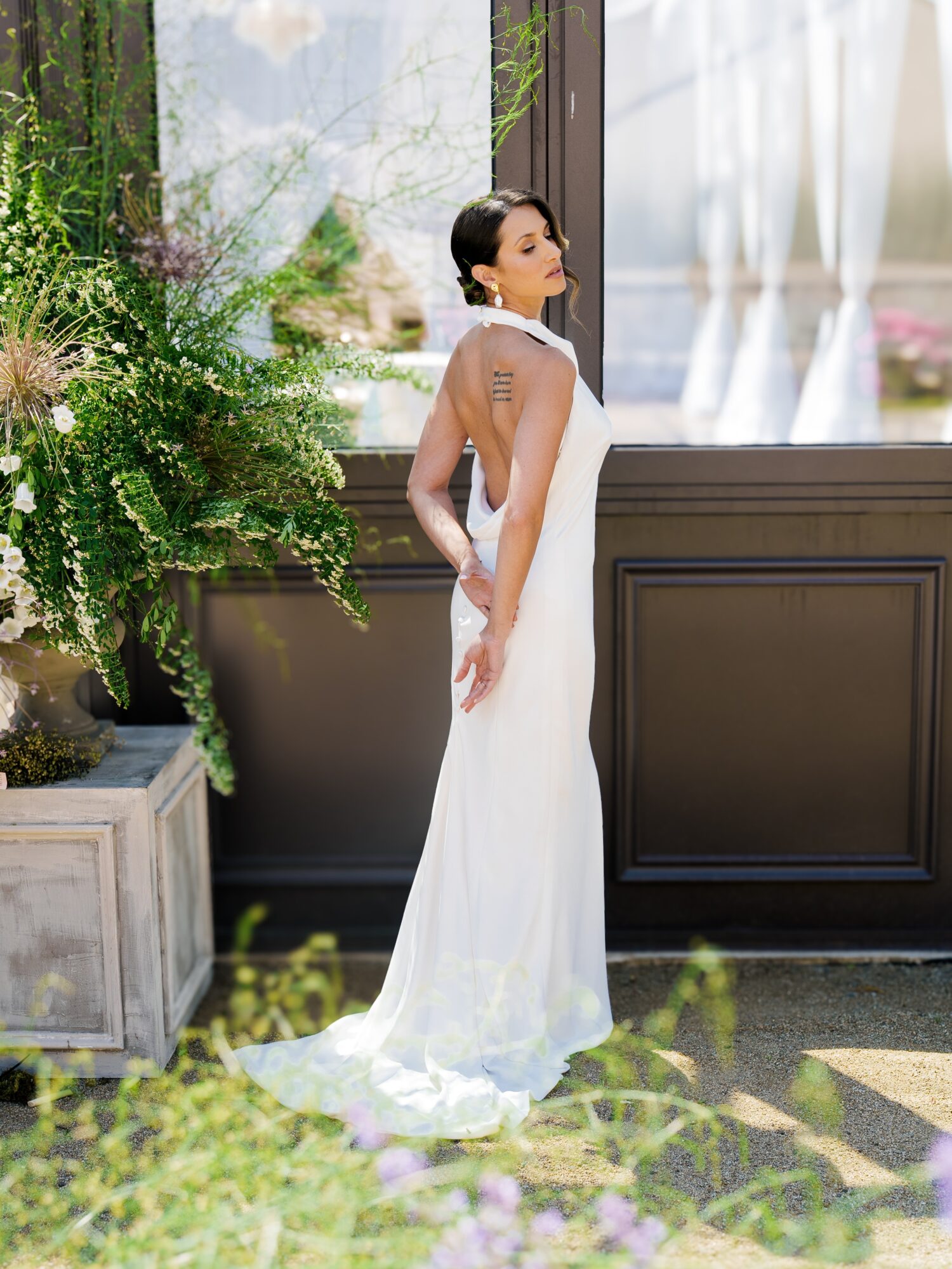 Woman in a white dress standing indoors near a window with plants, facing sideways, with sunlight streaming in.