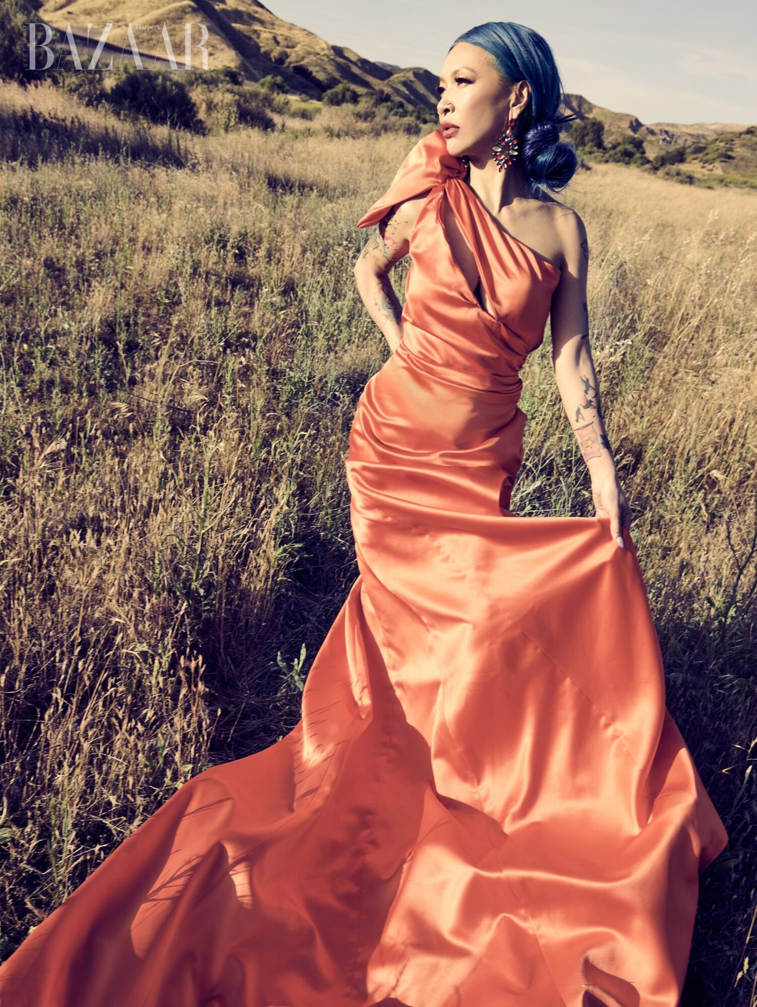 Woman in an orange satin gown standing in a grassy field with hills in the background.