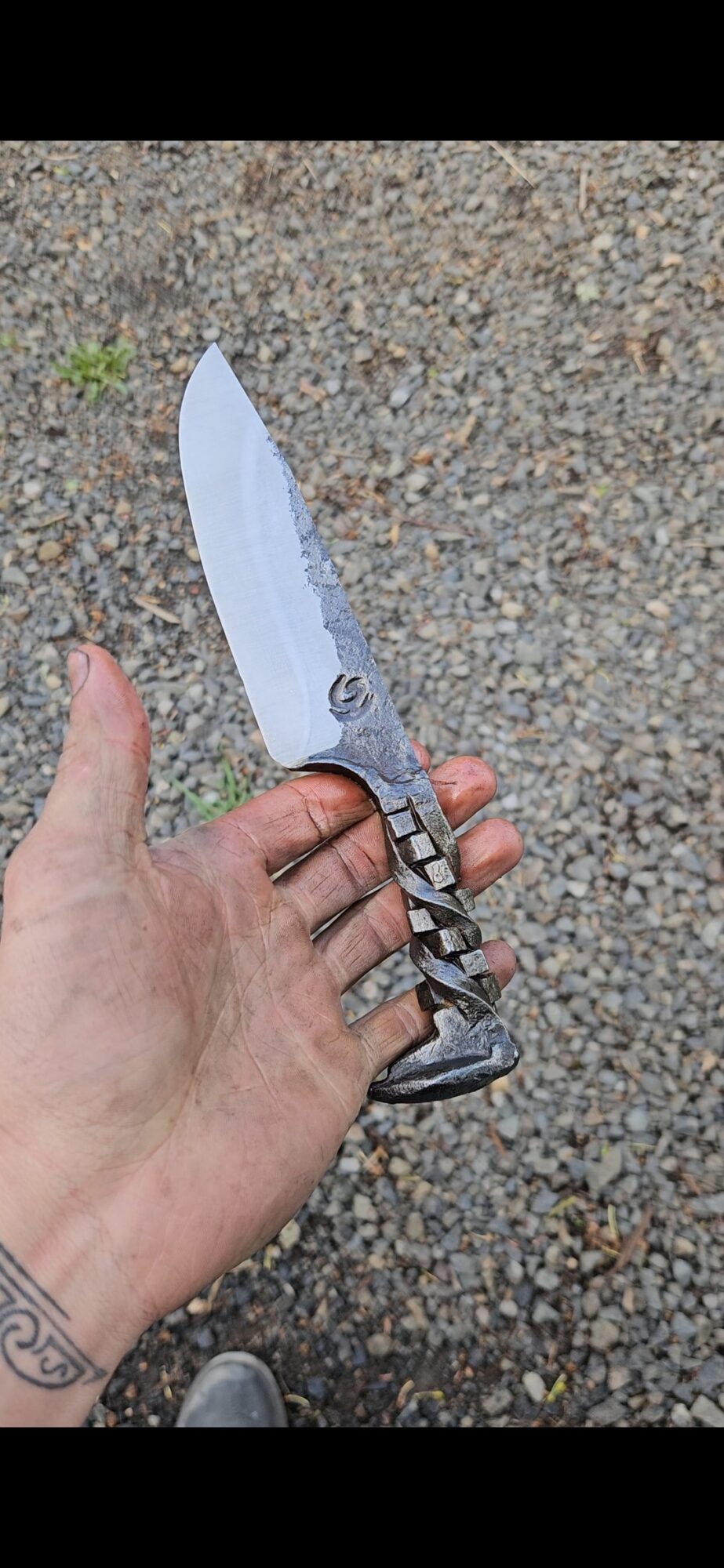 Hand holding a large knife with a patterned handle, outdoors on gravel ground.