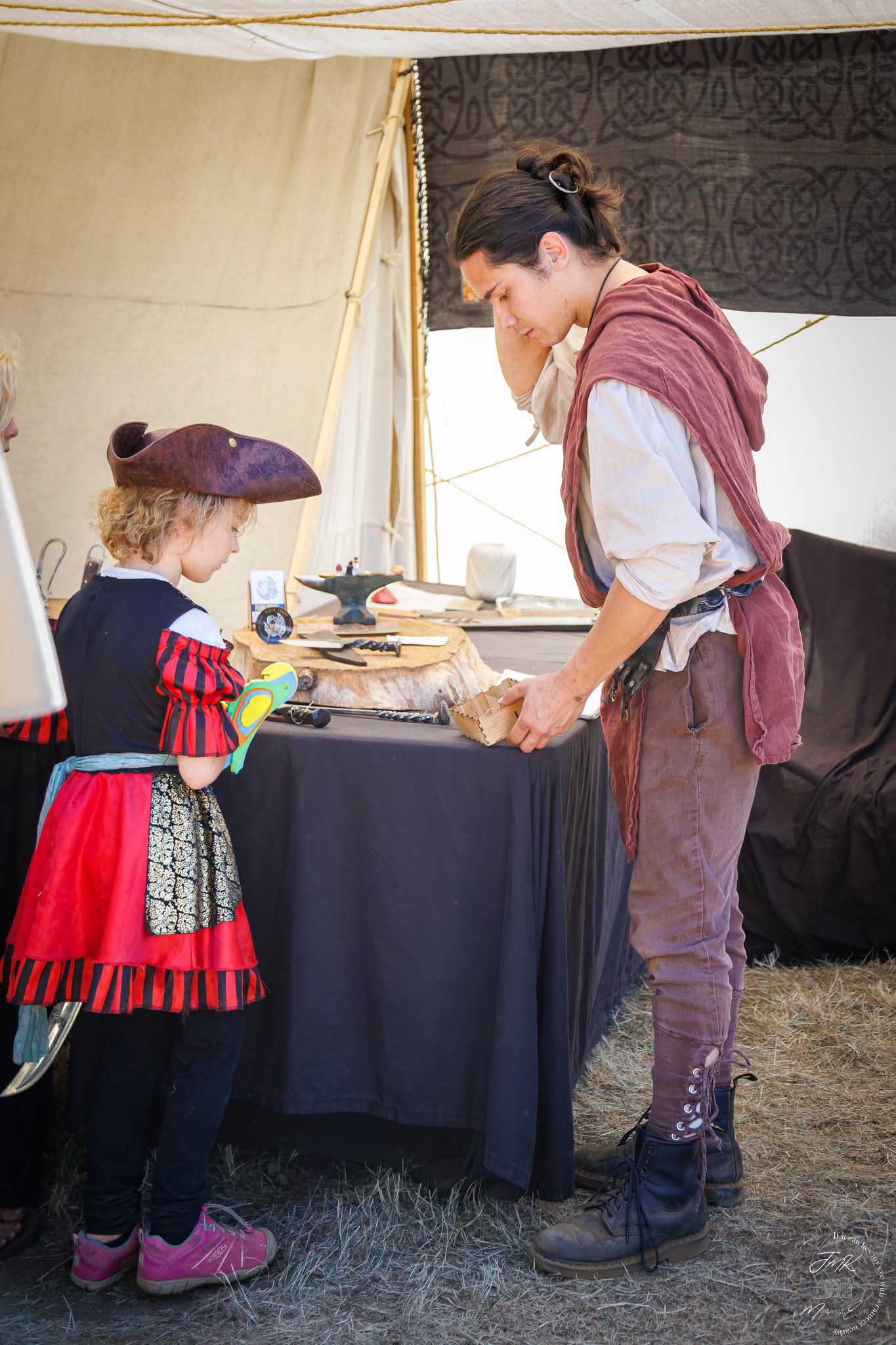 Two women in historical costumes at a booth with various items, one woman is showing something to the other.