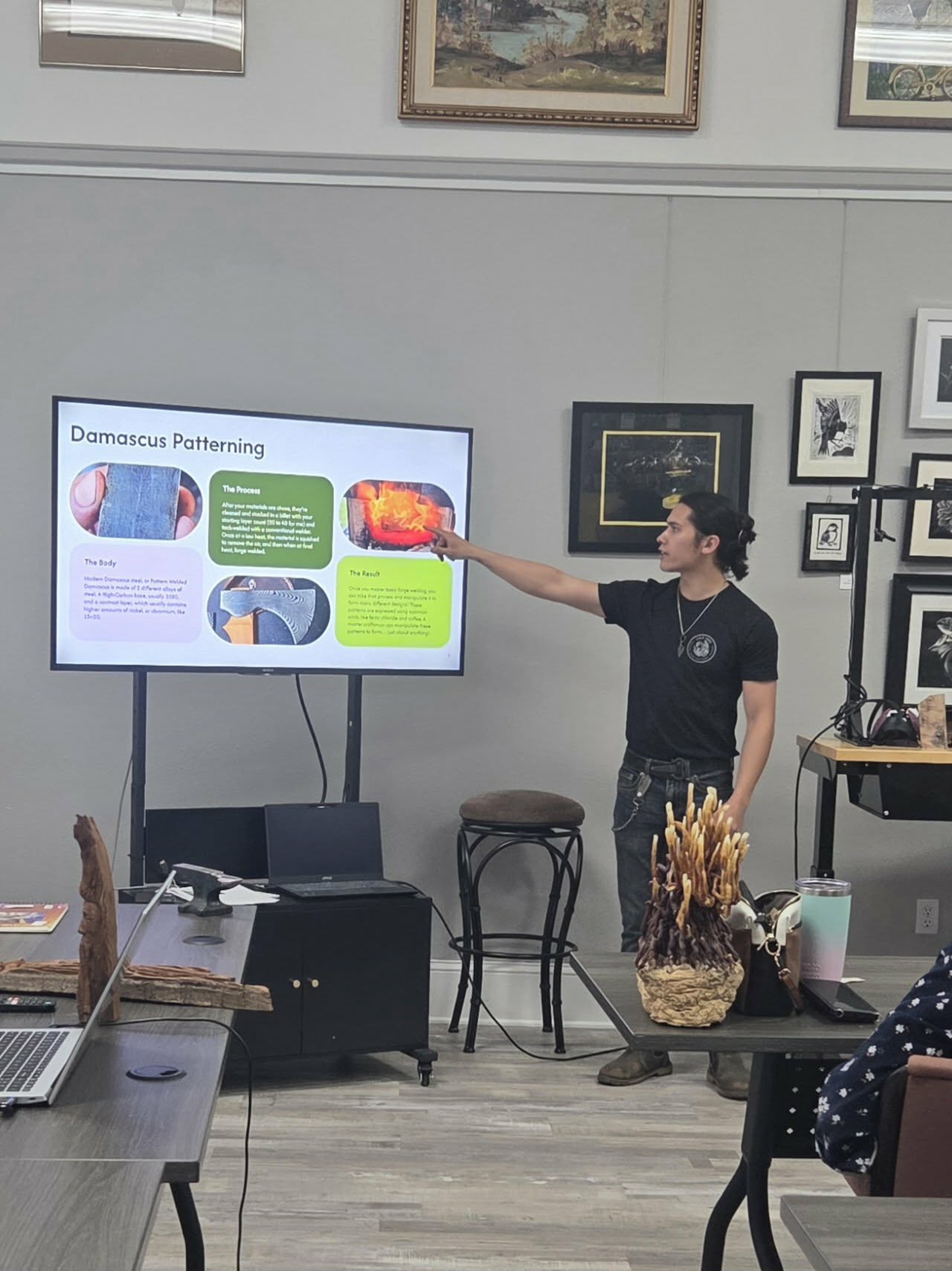 Person presenting a slide on a large screen in a room with framed pictures on the wall.