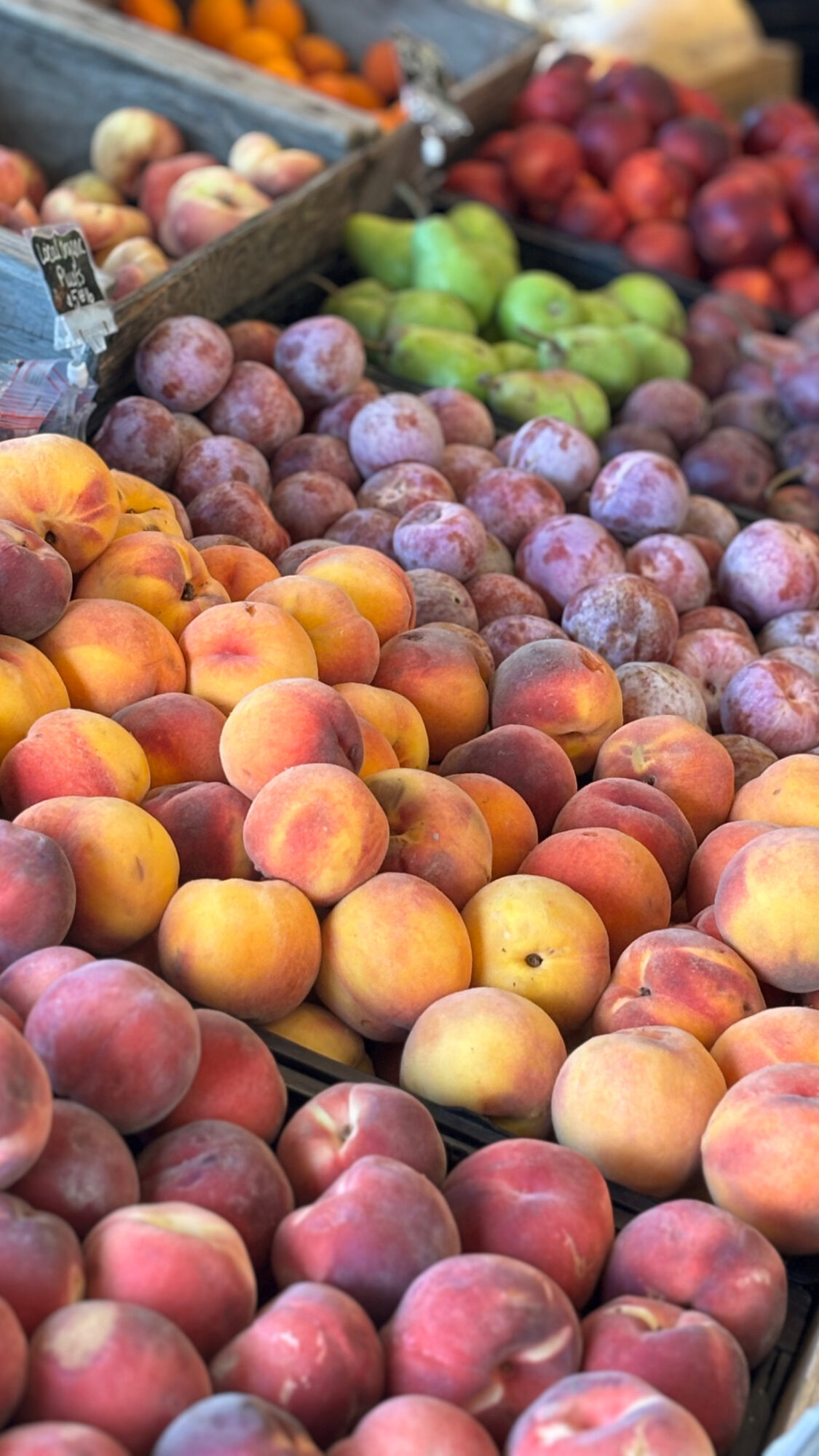 Display of various fruits including peaches, plums, apples, and onions arranged in sections at a market stall.