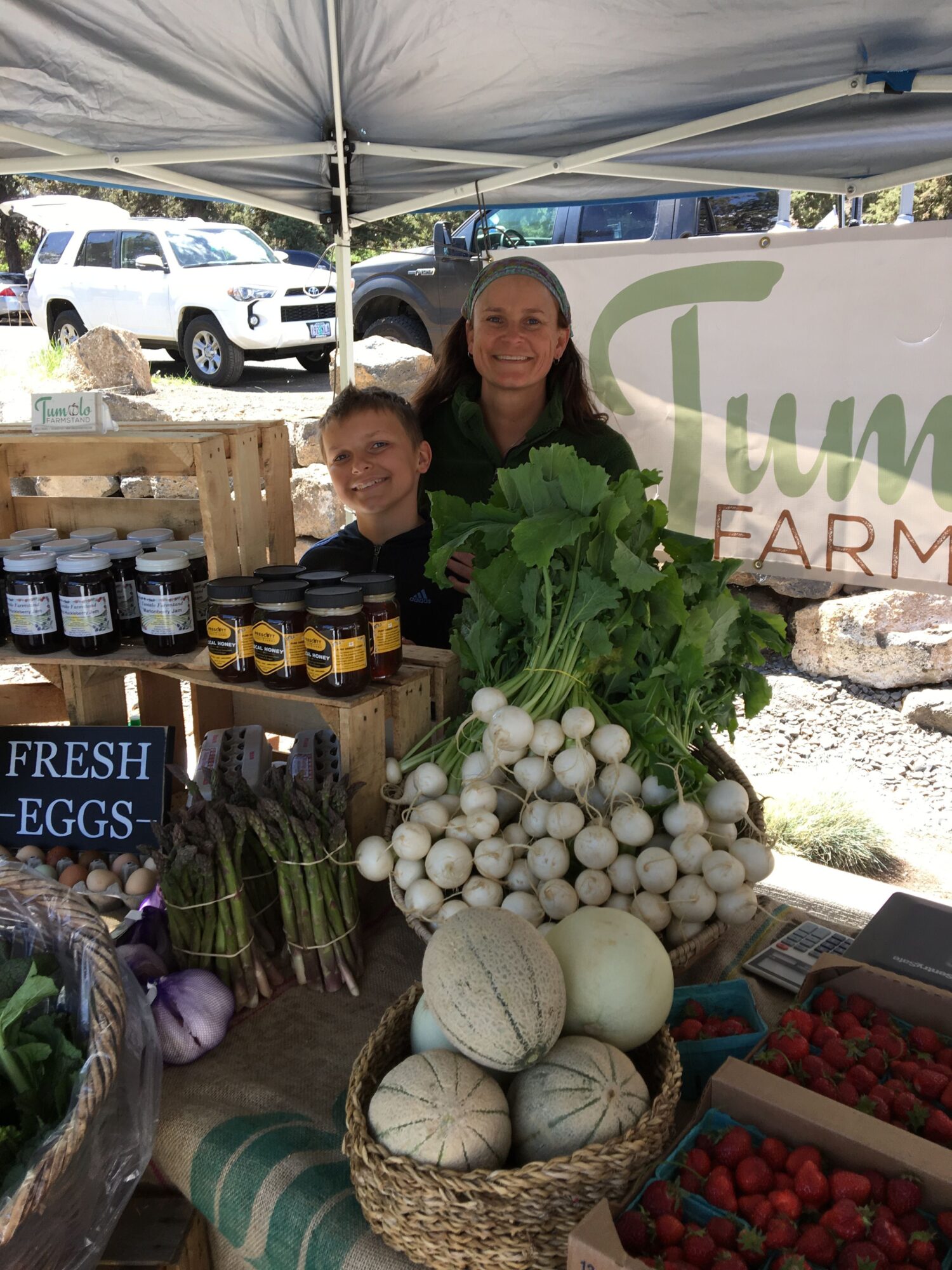 Two women stand behind a display of fresh vegetables including onions, garlic, and melons at an outdoor market stall.