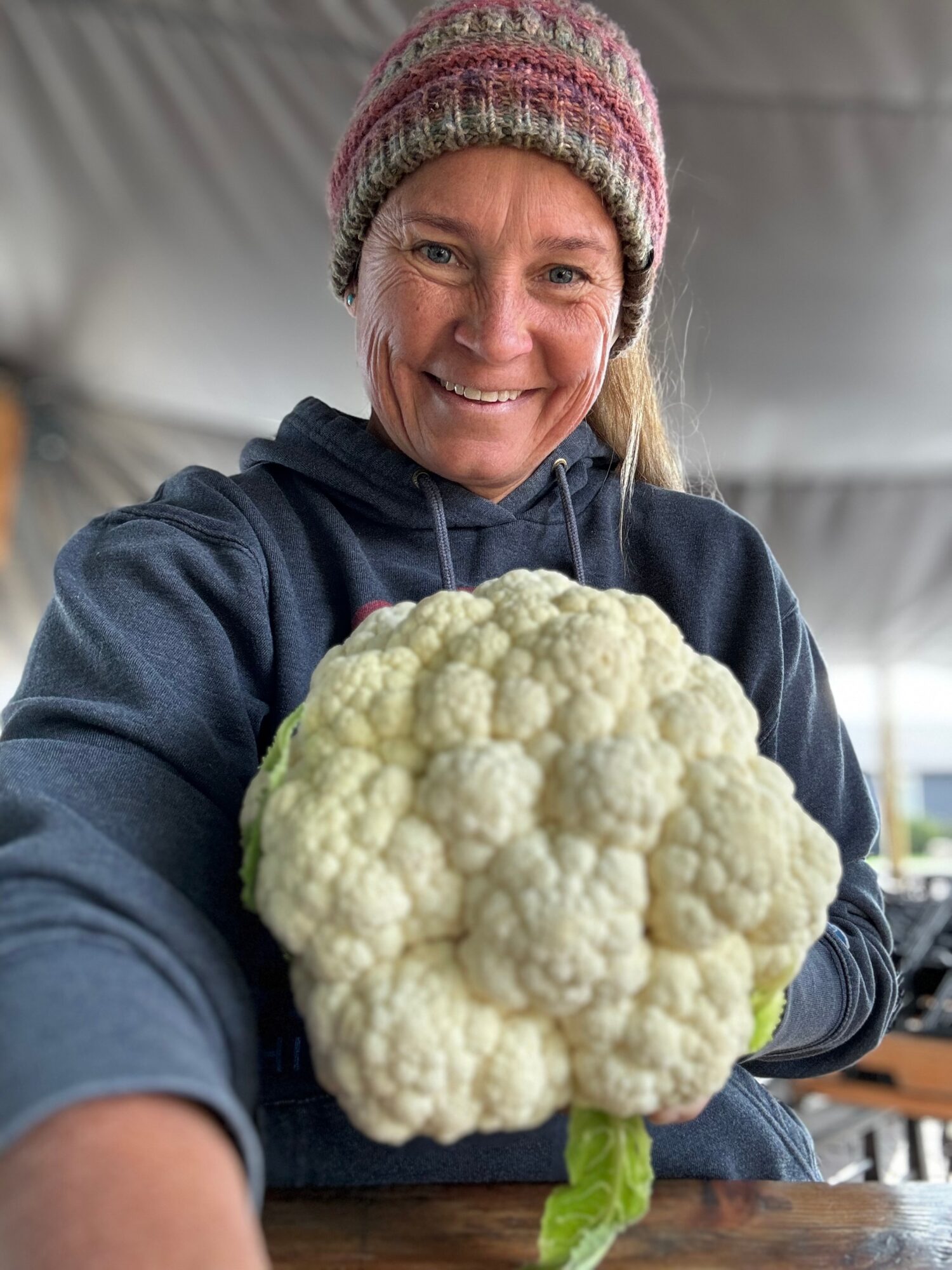 Woman in a pink knit hat and dark hoodie holding a large cauliflower, smiling, indoors with a tent-like ceiling.