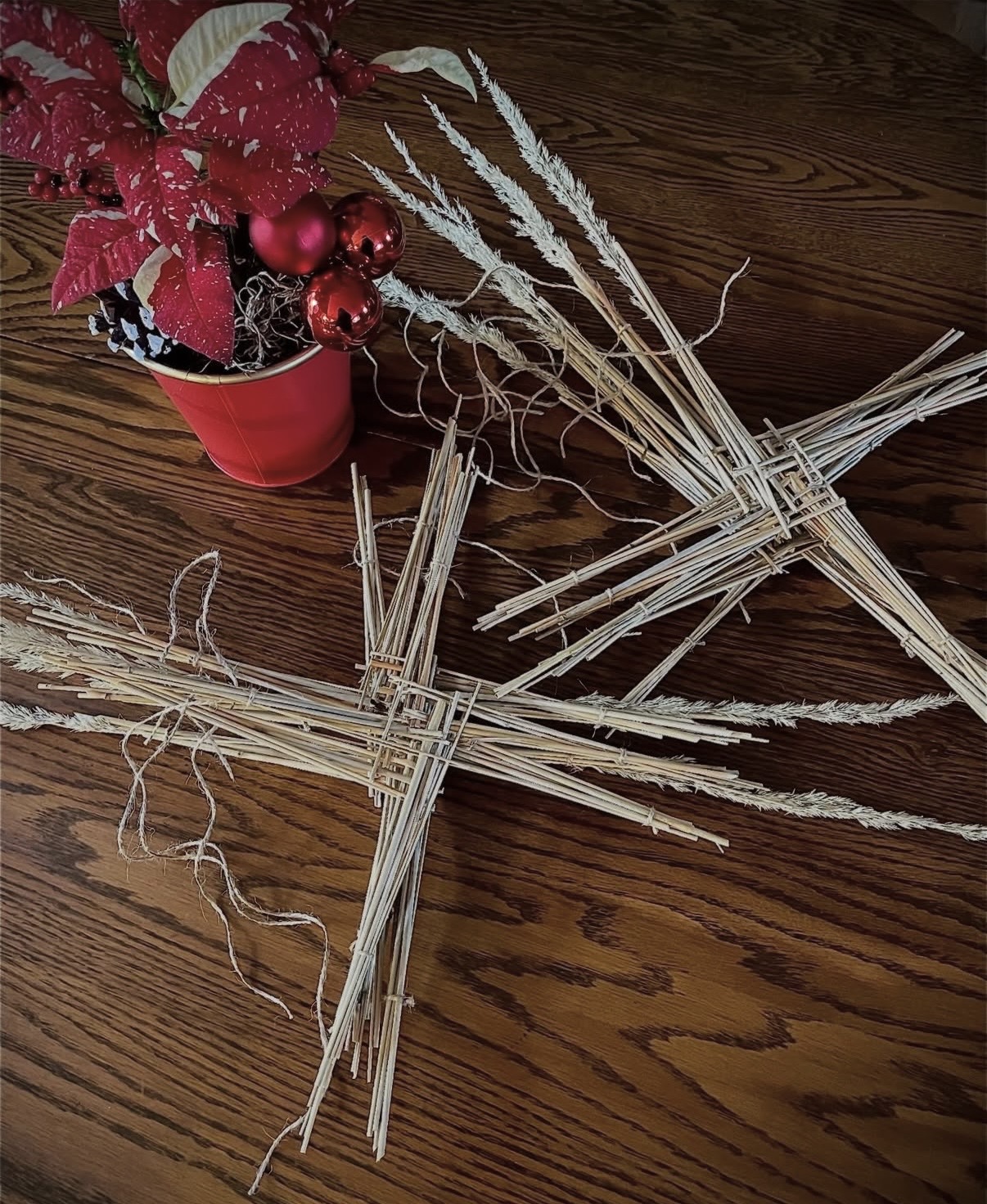 Decorative straw figures on a wooden surface with a red pot containing red and black ornaments and poinsettia flowers.