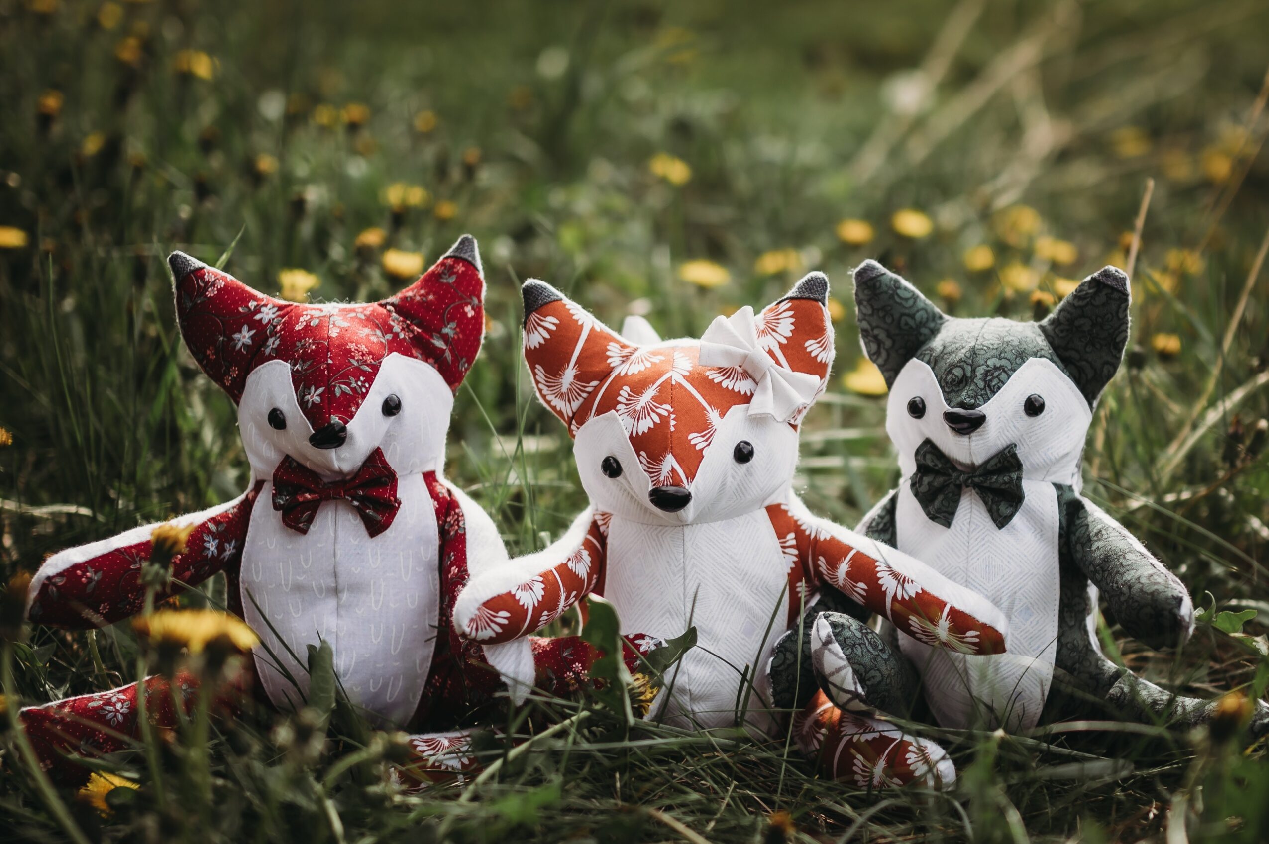 Three plush animal toys resembling foxes and cats sit on grass with yellow flowers, outdoors during daytime.