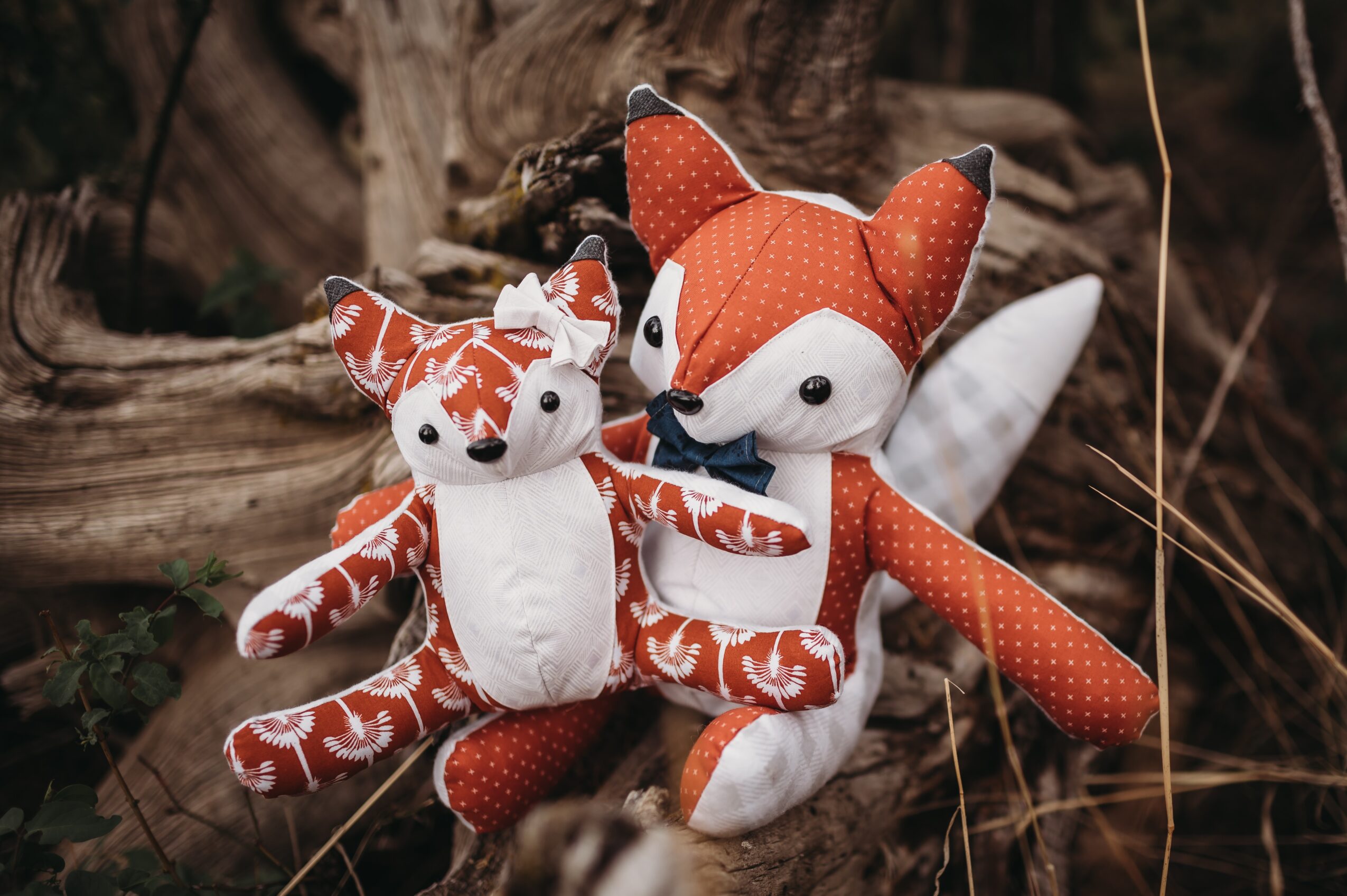 Two plush fox toys, one larger and one smaller, sitting on a tree trunk outdoors, surrounded by natural elements.